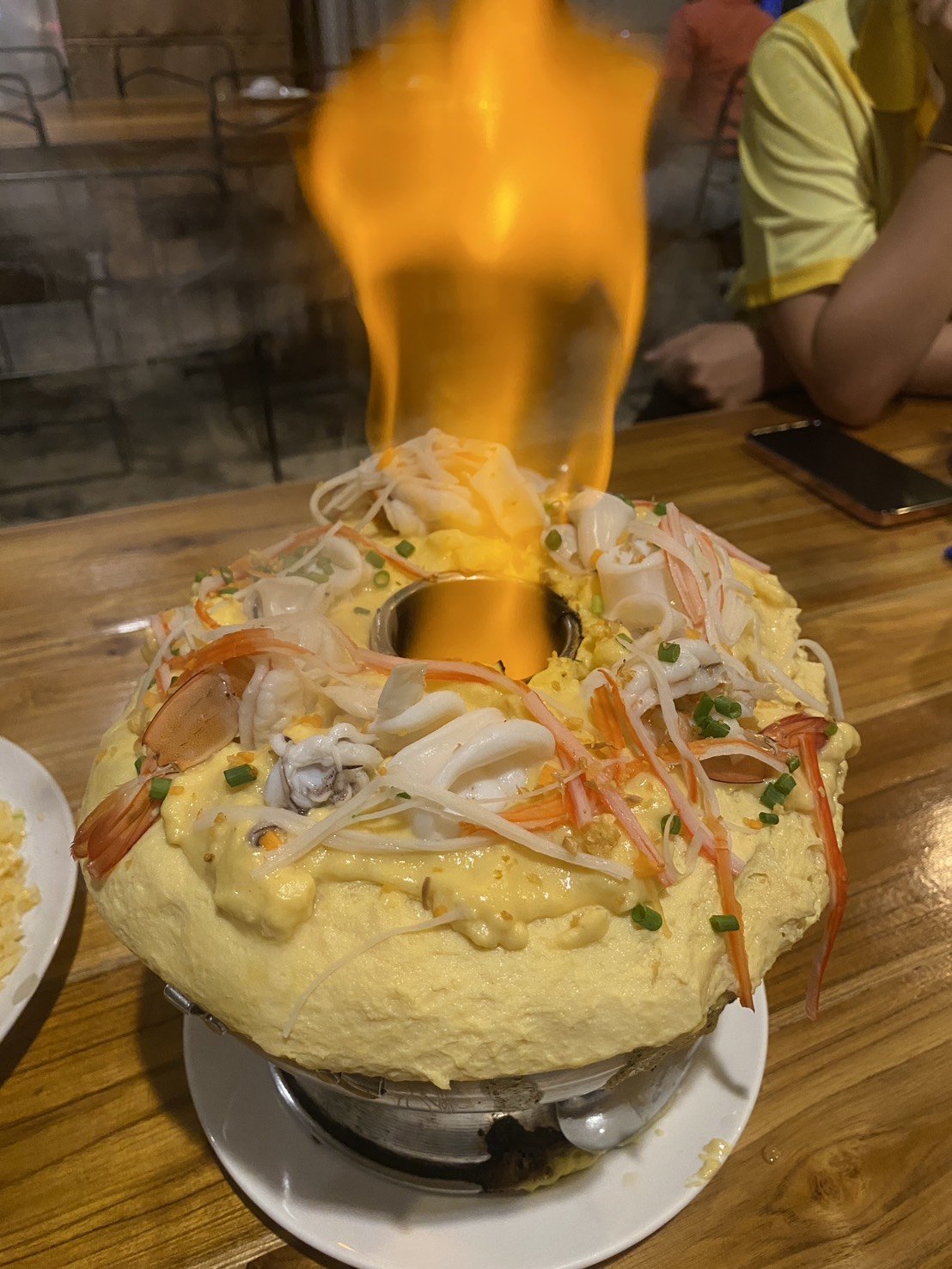 A seafood hot pot topped with a fluffy yellow egg omelet, seafood pieces, green onions, and a drizzle of sauce, with a flame above it. Kids English Thailand experience!
