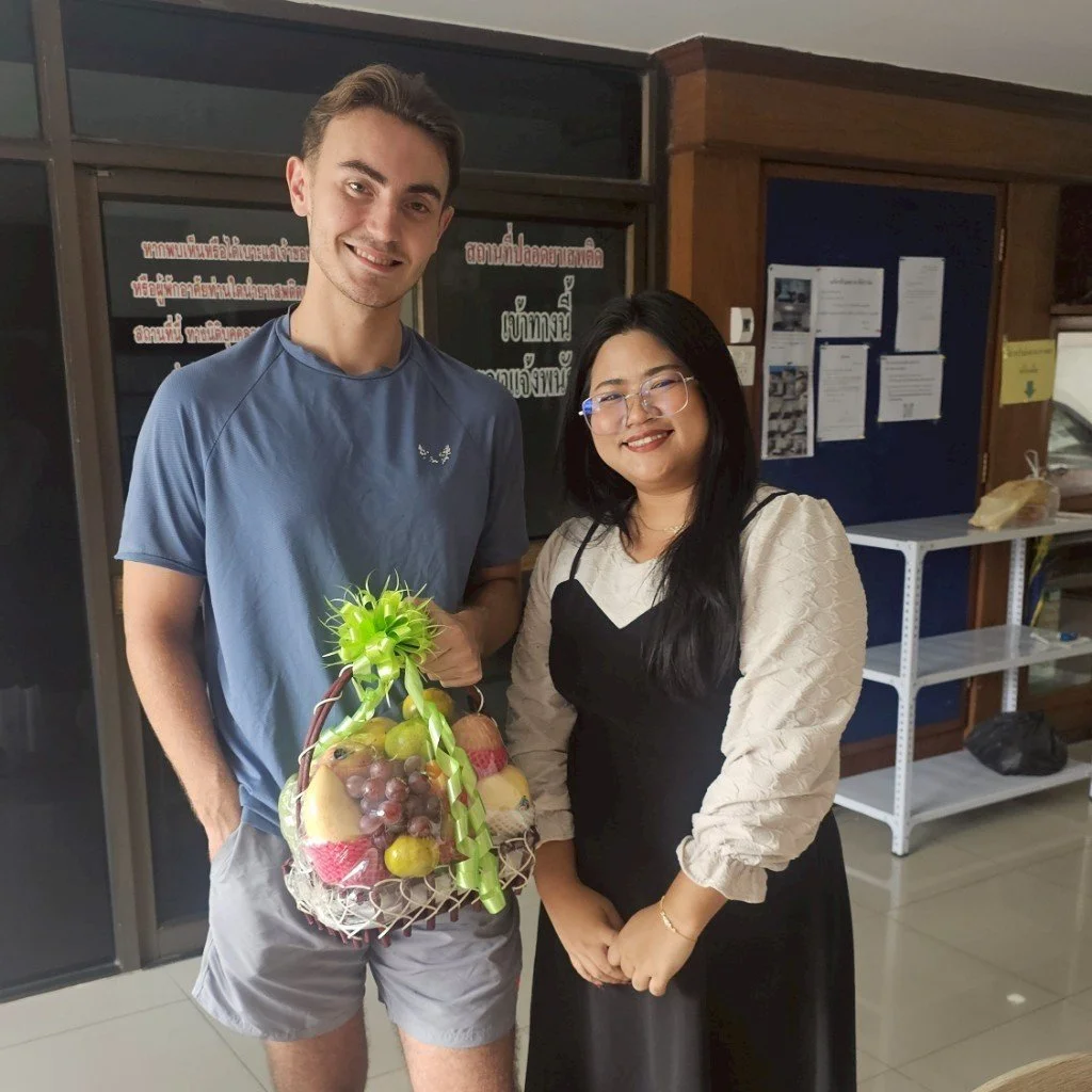 A young man and woman from Kids English Thailand standing indoors, smiling at the camera. The man is holding a fruit basket decorated with a green ribbon. The background features a dark board with notices and a white shelving unit.