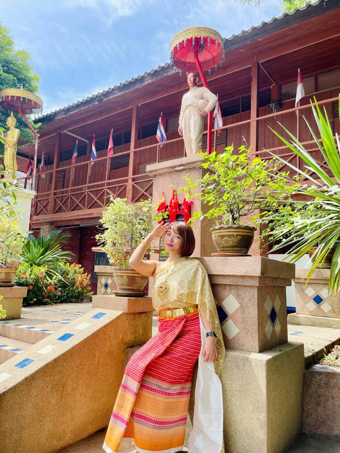 A Kids English Thailand teacher dressed in traditional Thai attire, posing outdoors in front of a statue and greenery, with a wooden building and flags in the background.