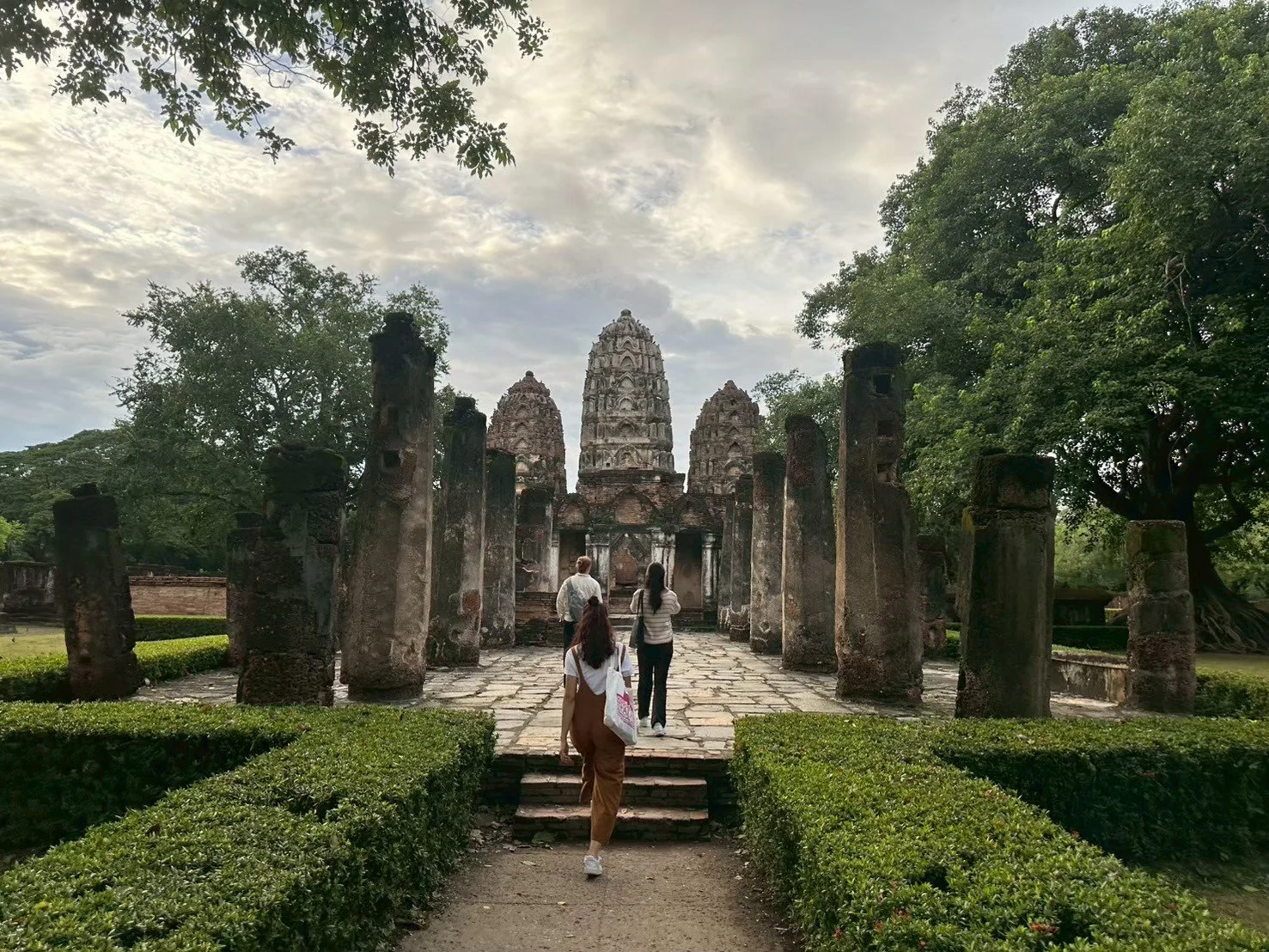 People from Kids English Thailand walking toward ancient stone temple ruins surrounded by greenery on a partly cloudy day.