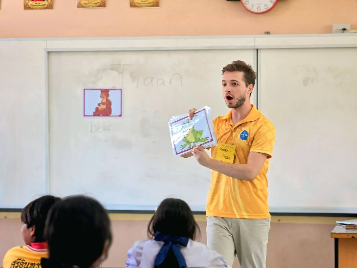 AKids English Thailand teacher in a yellow KET shirt holding flashcards with pictures of a bear and a frog in front of a classroom, with students seated facing him. The whiteboard behind has the words 'bear' and 'frog' written on it.