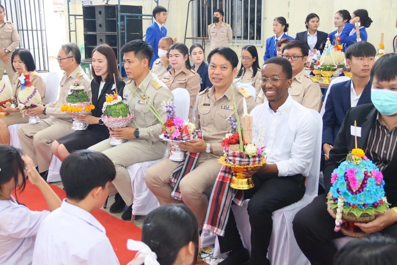 Group of Kids English Thailand teachers sitting and holding traditional floral arrangements in an indoor event.