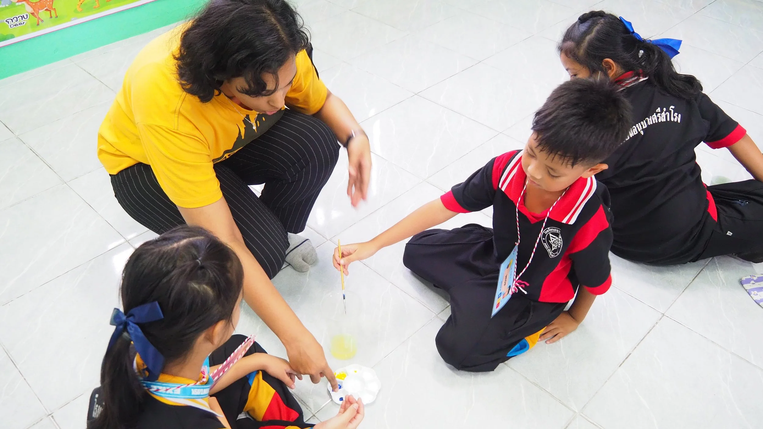 A Kids English Thailand teacher and three students sitting on the floor engaged in a science experiment or activity, with the teacher helping the children.