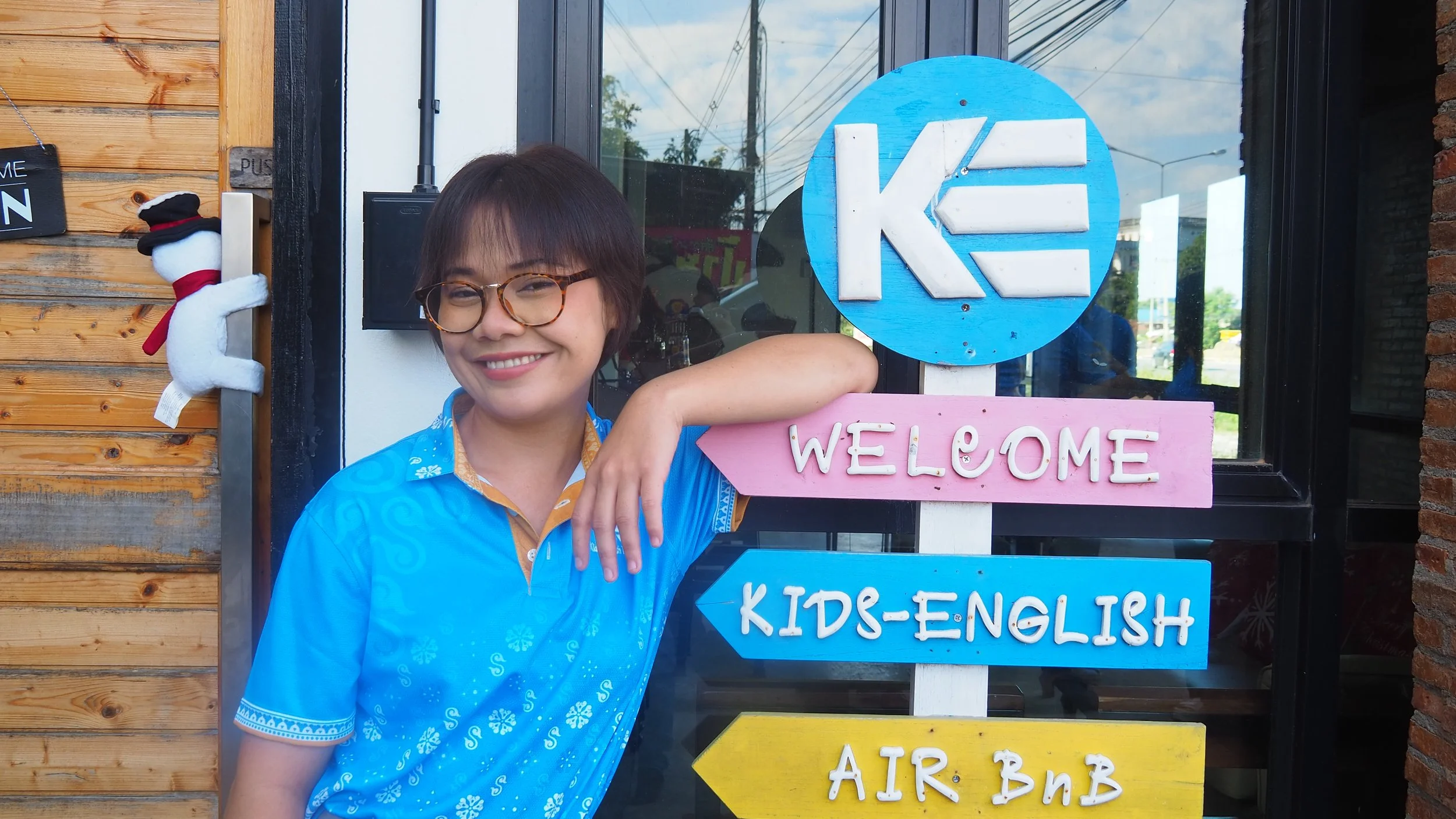 Woman holding a pink arrow sign that says 'Welcome' next to a sign with colorful sections, the top blue with a logo, and the bottom yellow with unreadable text, standing in front of a building with wooden siding and a large window.