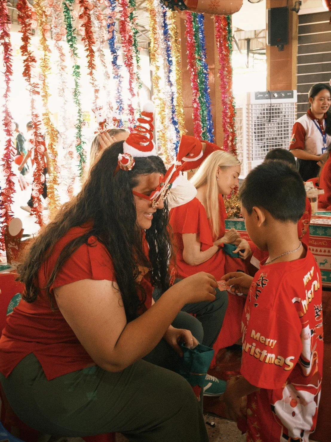 Kids English Thailand teachers celebrating Christmas with festive decorations, gift exchange, and holiday attire, including Christmas hats and shirts with holiday designs, in a decorated indoor space.