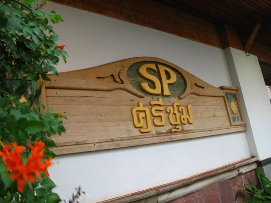 Wooden sign with gold lettering that reads 'SP' and some Asian script below it, mounted on a white wall with greenery and orange flowers nearby.