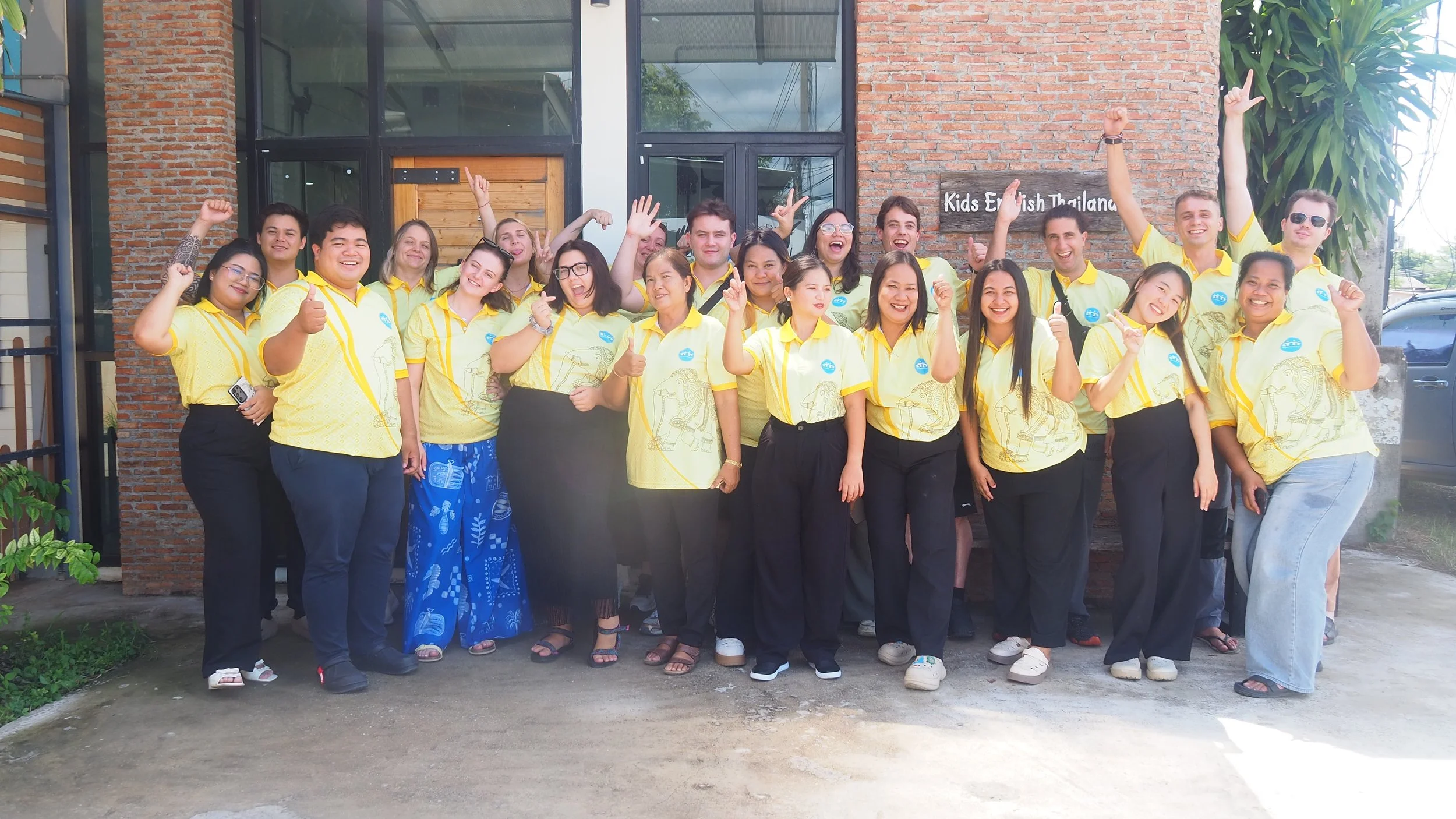 Group of smiling Kids English Thailand teachers wearing yellow shirts standing outside a building with a sign that says "Kids English Thailand". Some people are raising their hands, giving thumbs up, or making peace signs.