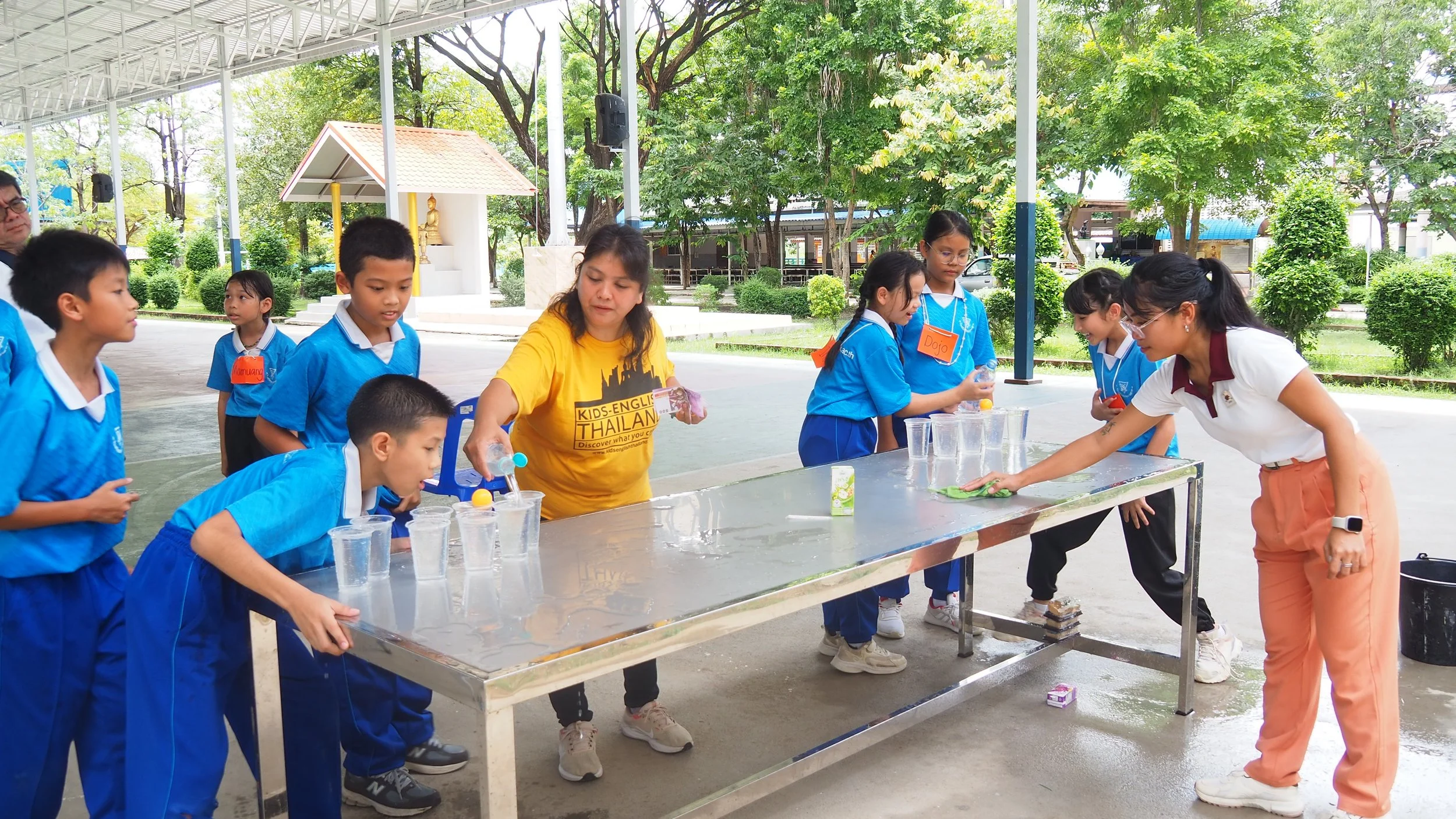 Children and Kids English Thailand teachers participate in an outdoor water activity under a covered area, with trees and greenery in the background.