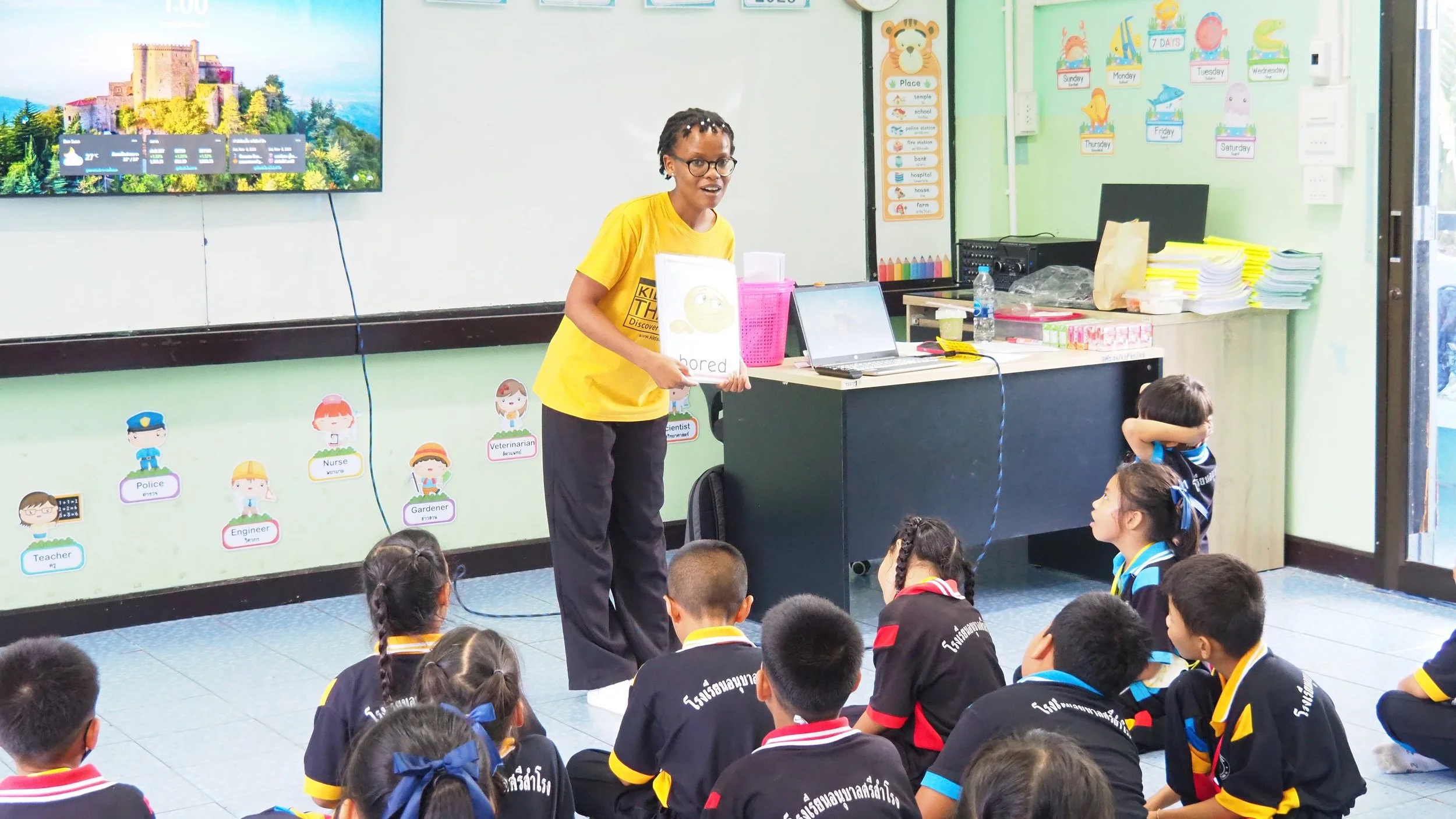 A Kids English Thailand teacher in a yellow shirt standing in front of a classroom of young students. The teacher is holding a whiteboard with the word 'bored' and a sad face drawing. The students are seated on the floor, listening attentively.