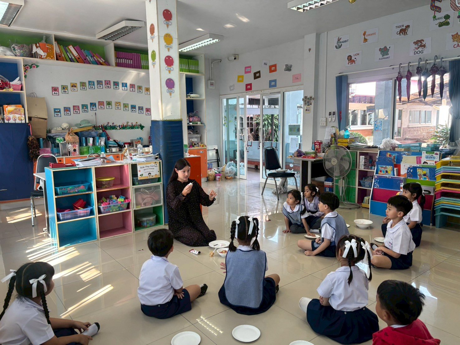 A teacher from Kids English Thailand is sitting on the floor and talking to a group of young children in a classroom. The children are sitting cross-legged and listening attentively. 