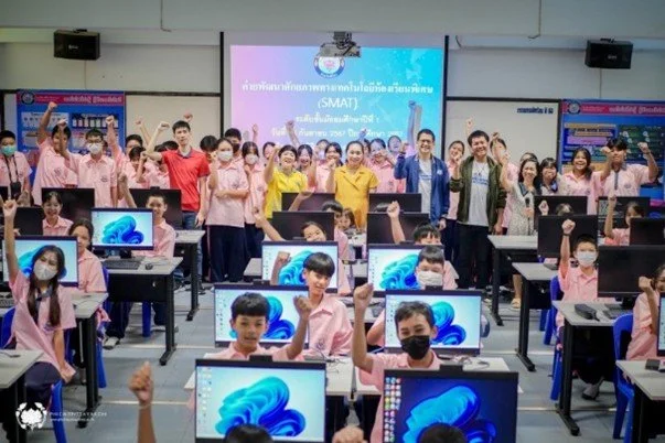 Kids English Thailand Image-School computer room with students and teachers raising hands, seated at computers with Windows desktops, large screen at back showing presentation in Thai language.