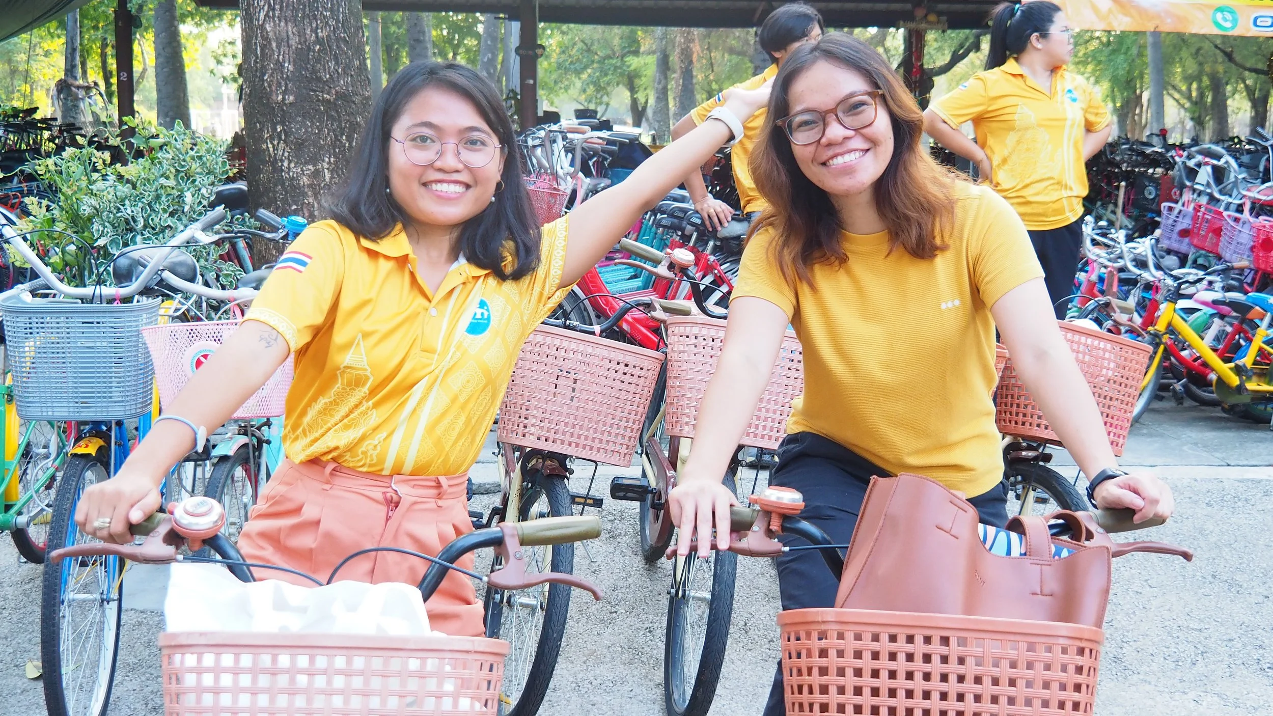 Two Kids English Thailand teachers wearing yellow shirts smiling and riding bicycles with front baskets, with numerous bicycles and trees in the background.