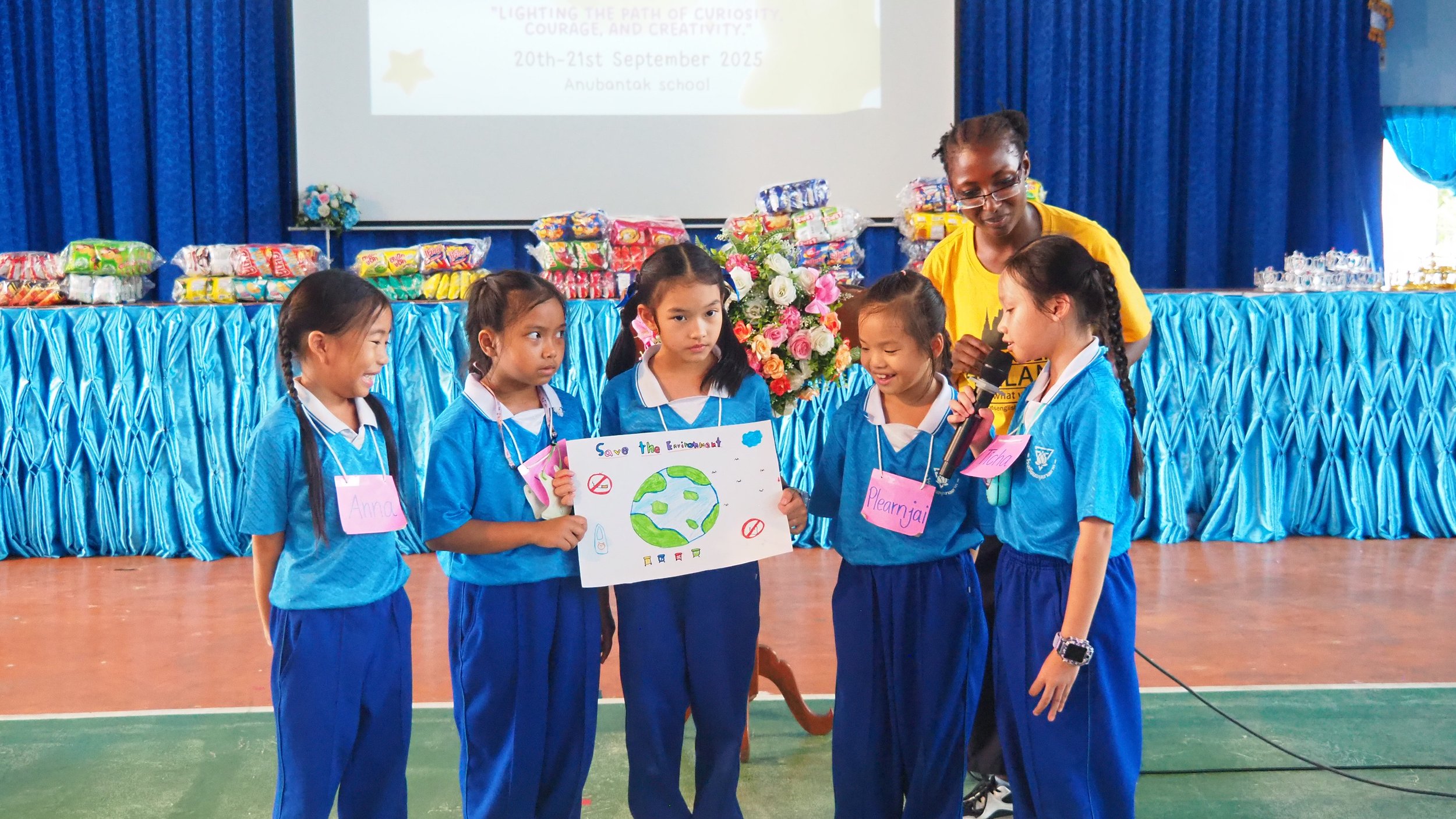 Five young girls in blue school uniforms with pink name tags, standing on a gymnasium floor, with a Kids English Thailand teacher participating in an environmental awareness presentation. 