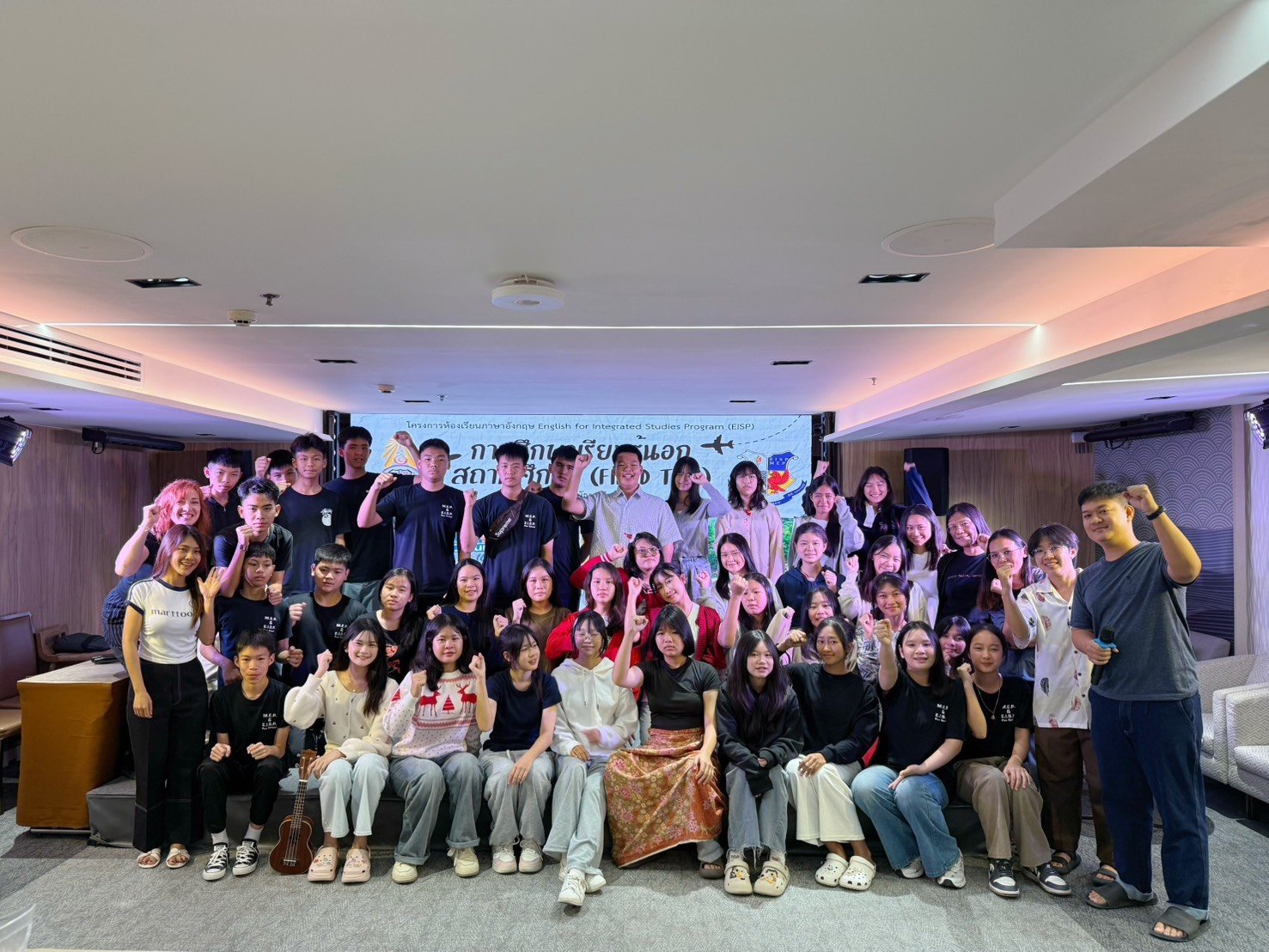 Group of young people and Kids English Thailand teachers posing for a photo during an event, with a banner in the background indicating the event is related to an English language program.