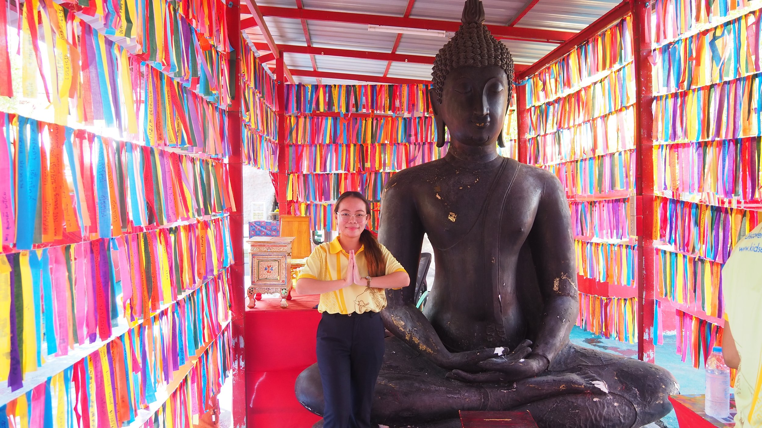 A KET Teacher in a yellow shirt with a prayer gesture and glasses standing next to a large, seated Buddha statue placed in a colorful, ribbon-lined shrine.