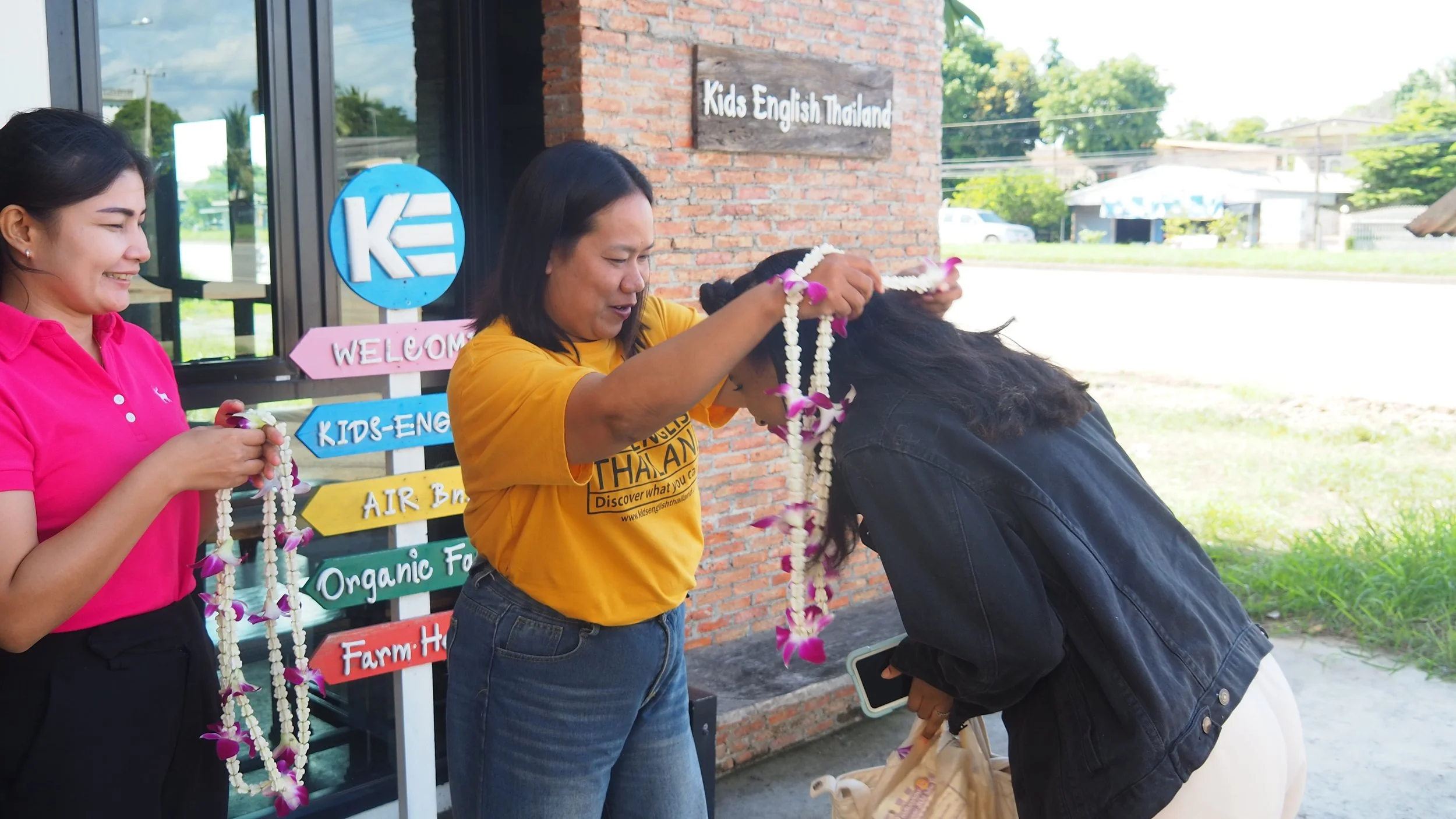 A Kids English Thailand team member in a yellow shirt drapes a flower garland over a Kids English Thailand teachers shoulders as part of a welcoming ceremony outside a building with a sign that reads 'Kids English Thailand'. 