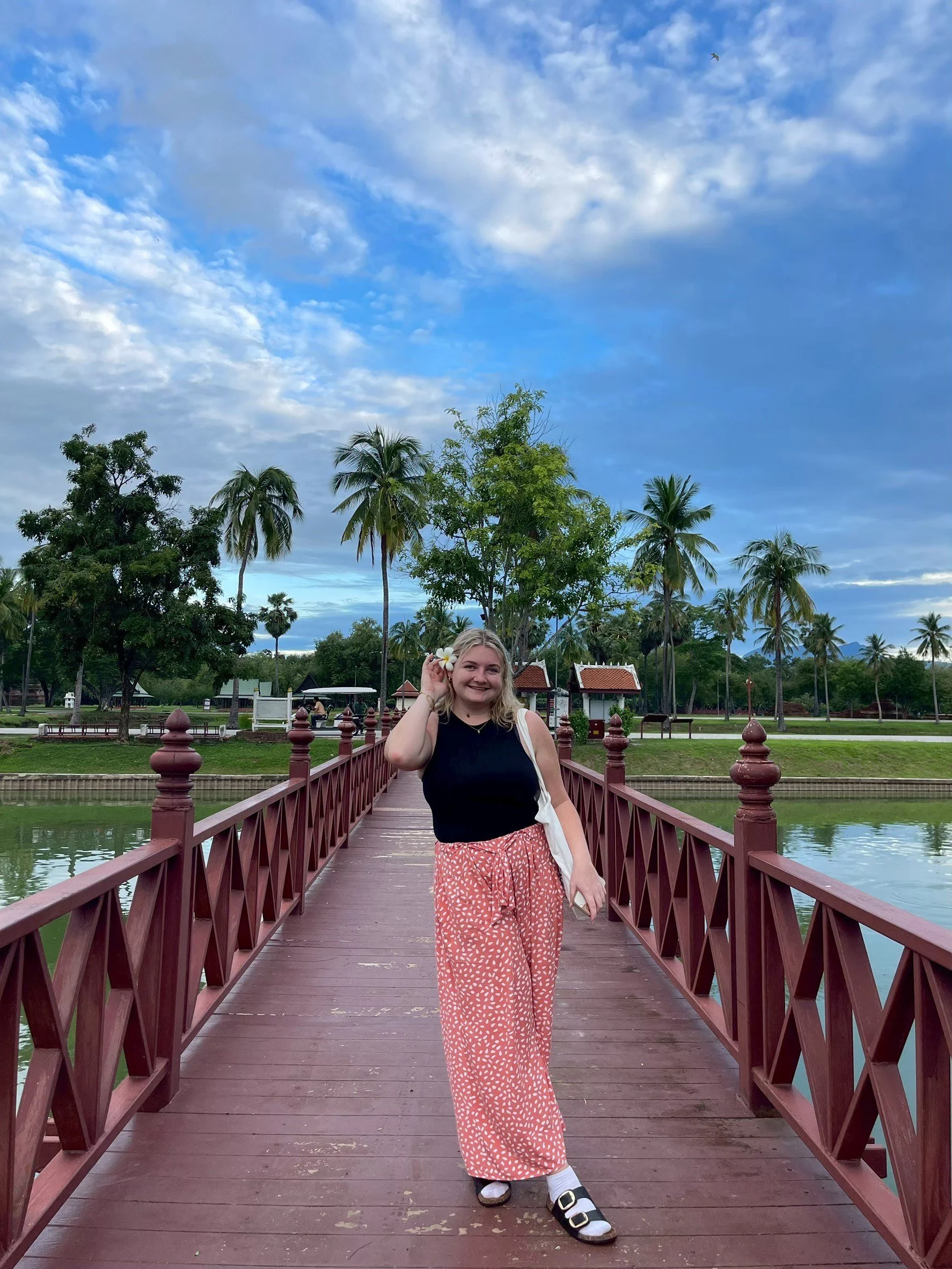 A smiling Kids English Thailand teacher standing on a wooden bridge over water in a park with palm trees, lush greenery, and a partly cloudy sky.