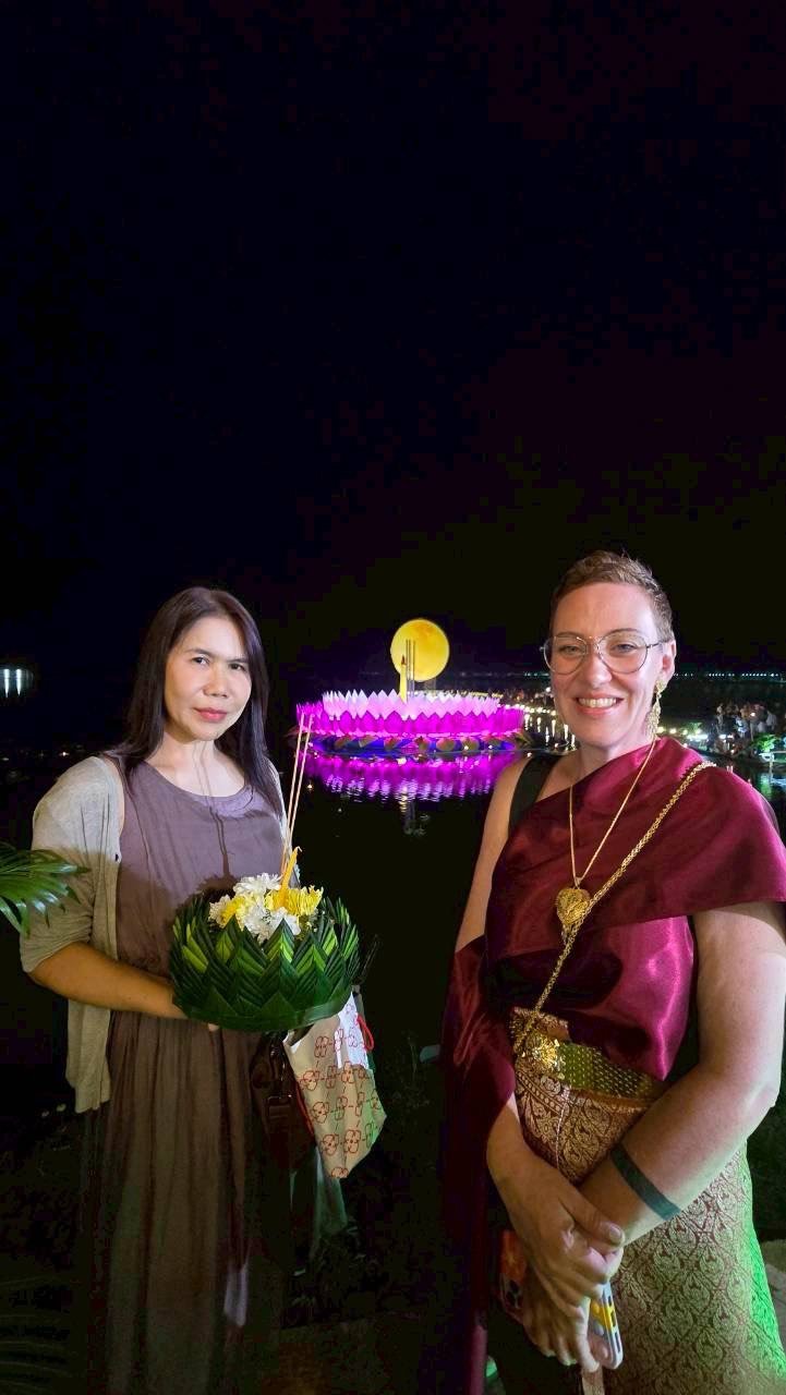 Two Kids English Thailand teachers standing outdoors at night with a colorful illuminated floating temple and lantern in the background.