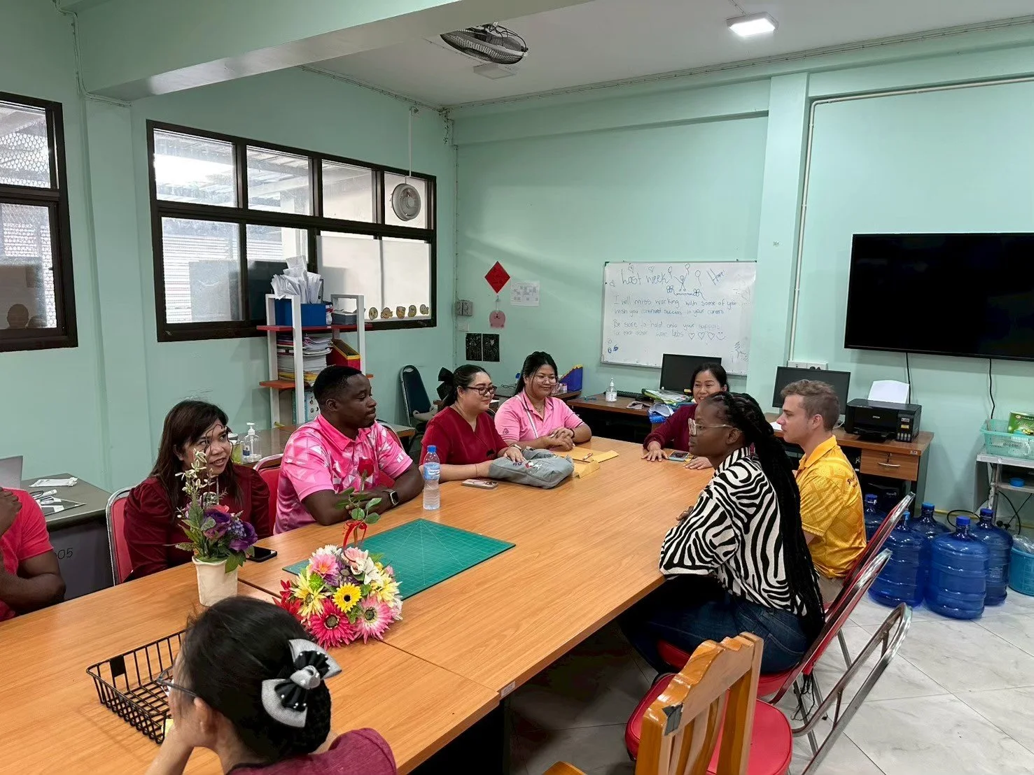 People from Kids English Thailand sitting around a conference table in a meeting room with light green walls, windows, and a large flat screen TV, engaged in a discussion.