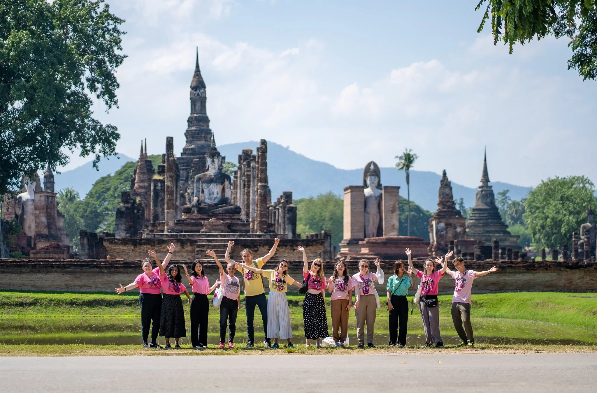 Group of Kids English Thailand teachers standing in front of ancient temple ruins with Buddha statues and mountains in the background, smiling and raising their hands.