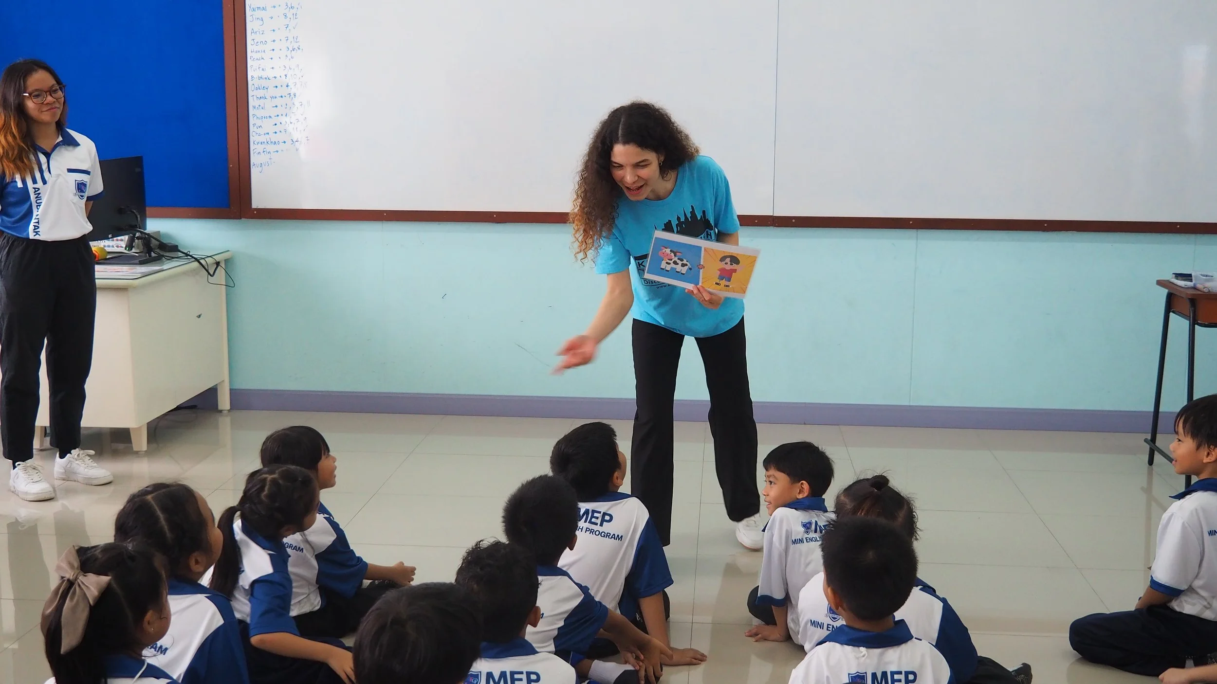 A classroom scene with a Kids English Thailand teacher in a blue KET shirt standing and reading to a seated group of young students dressed in white and blue uniforms. One student stands near a whiteboard on the left side of the image.