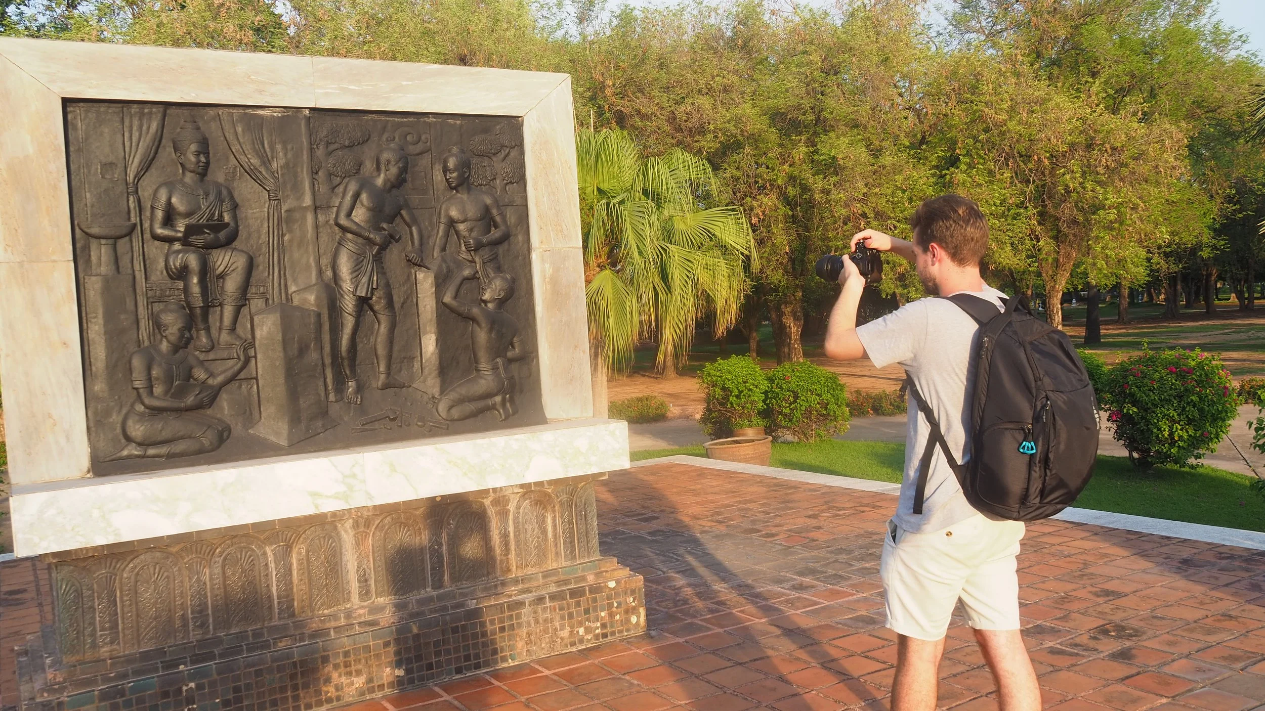 A Kids English Thailand teacher with a backpack taking a photograph of a bronze memorial sculpture in a park during sunset.
