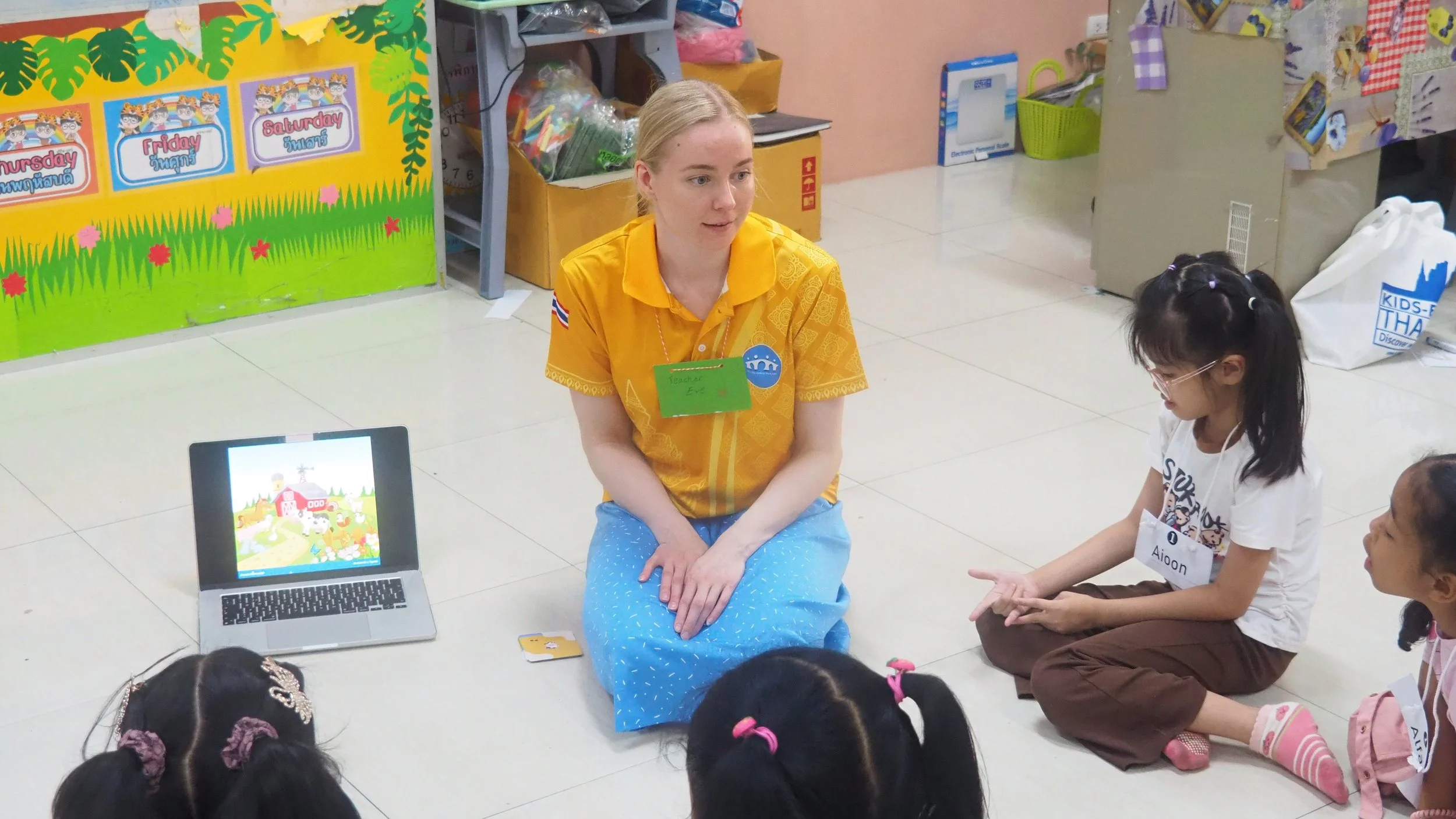 A Kids English Thailand teacher and young children sitting on the floor in a classroom, engaging in a lesson with a laptop and a colorful picture of a cartoon landscape on the screen.
