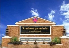 Kids English Thailand Image-A brick school building with a sign in front that reads 'World-Class Standard School' and a logo on the roof, under a blue sky with clouds.