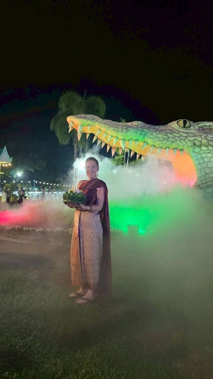 A Kids English Thailand teacher wearing traditional Southeast Asian attire holding a tray of offerings, standing in front of a large illuminated crocodile sculpture with colorful lights at night.