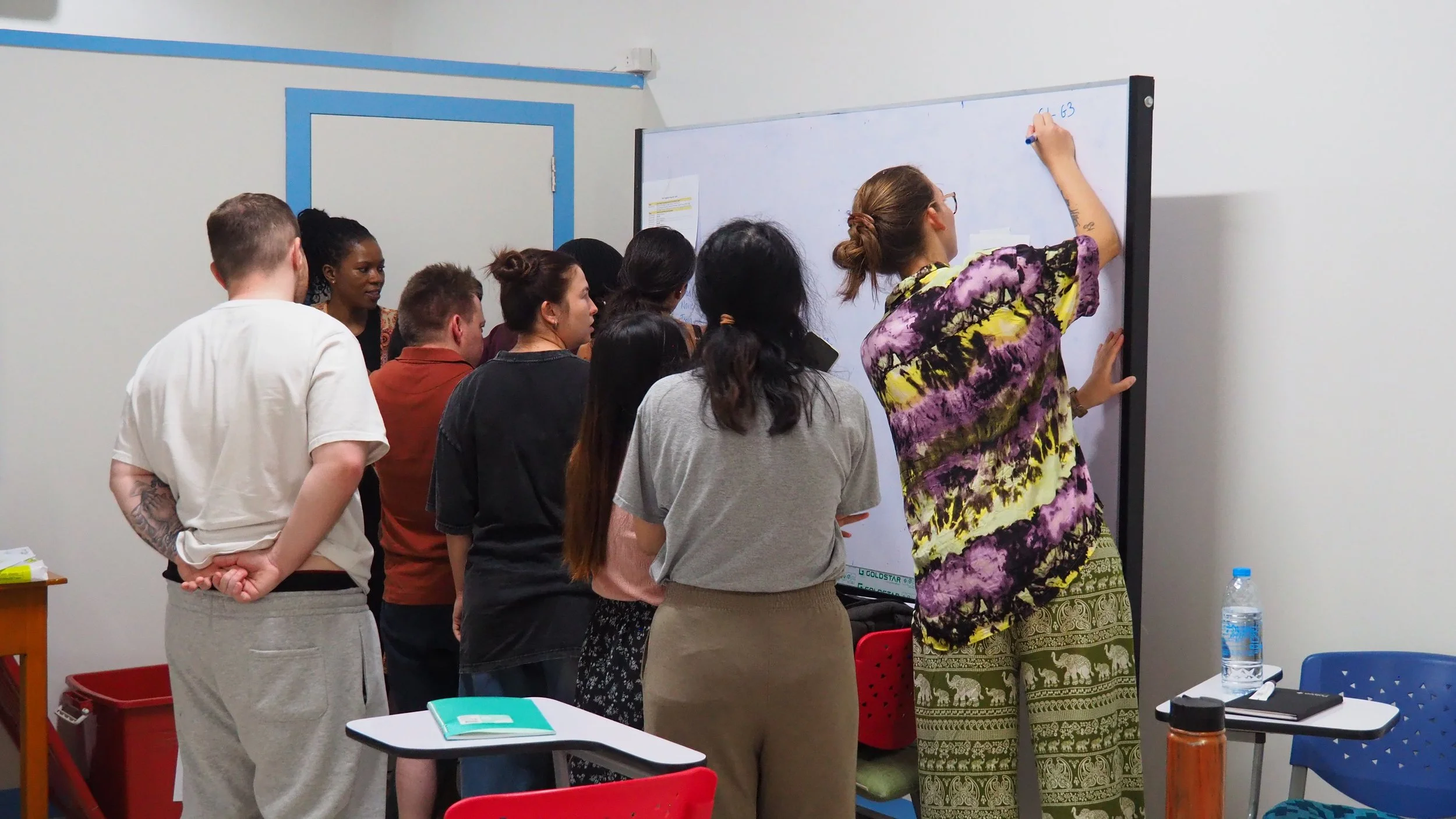 A group of Kids English Thailand teachers standing around a woman writing on a whiteboard in a classroom or meeting room.