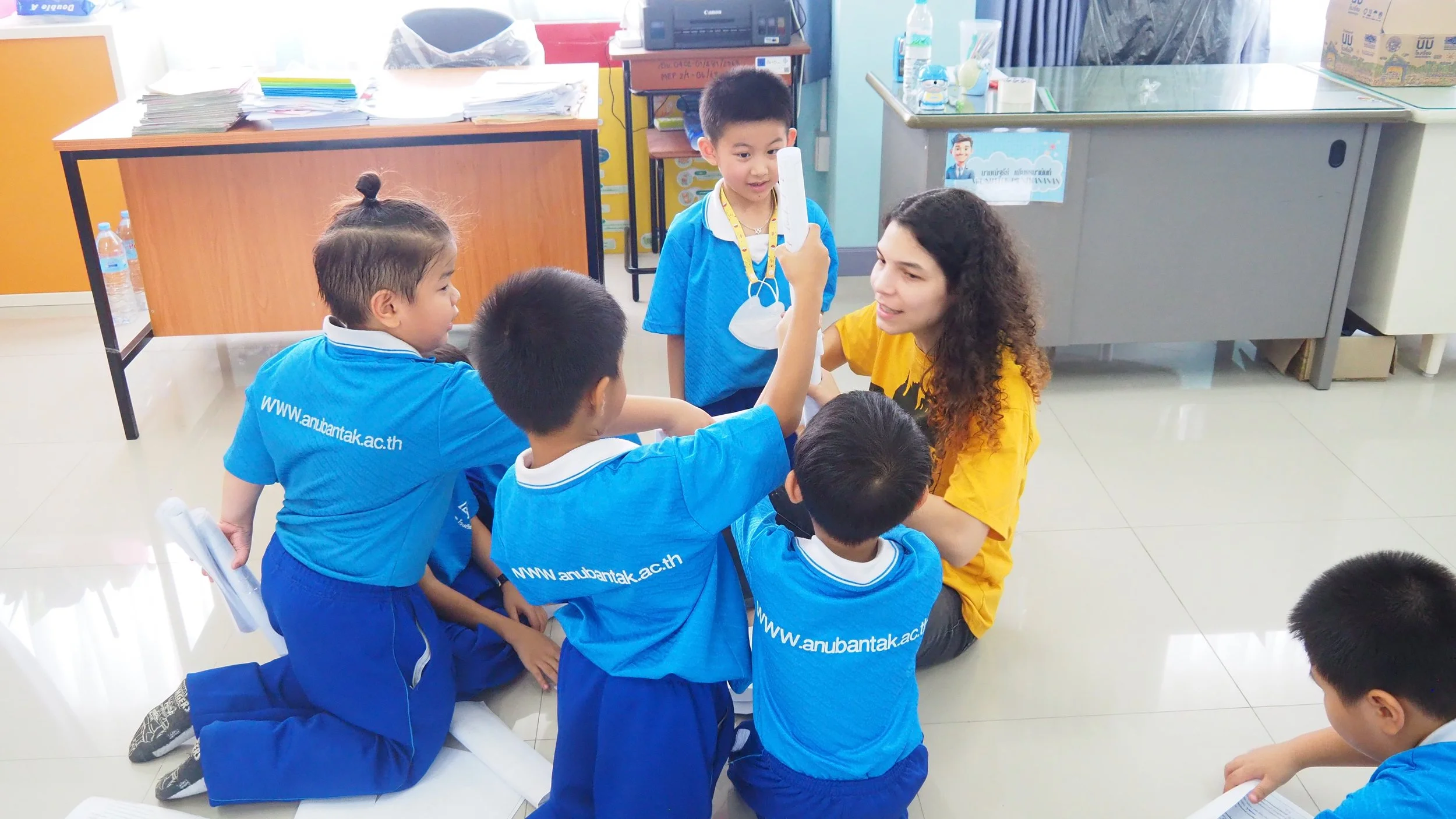 A Kids English Thailand teacher with curly hair and a yellow KET shirt interacting with five young students in blue uniforms sitting or kneeling on the floor in a classroom. 