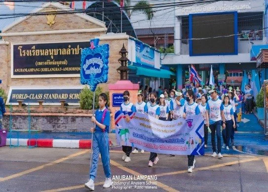Kids English Thailand Image-Students march in a parade outside a school with signs and banners, some wearing blue and white uniforms, in front of a sign for Anuban Lampang School in Thailand.