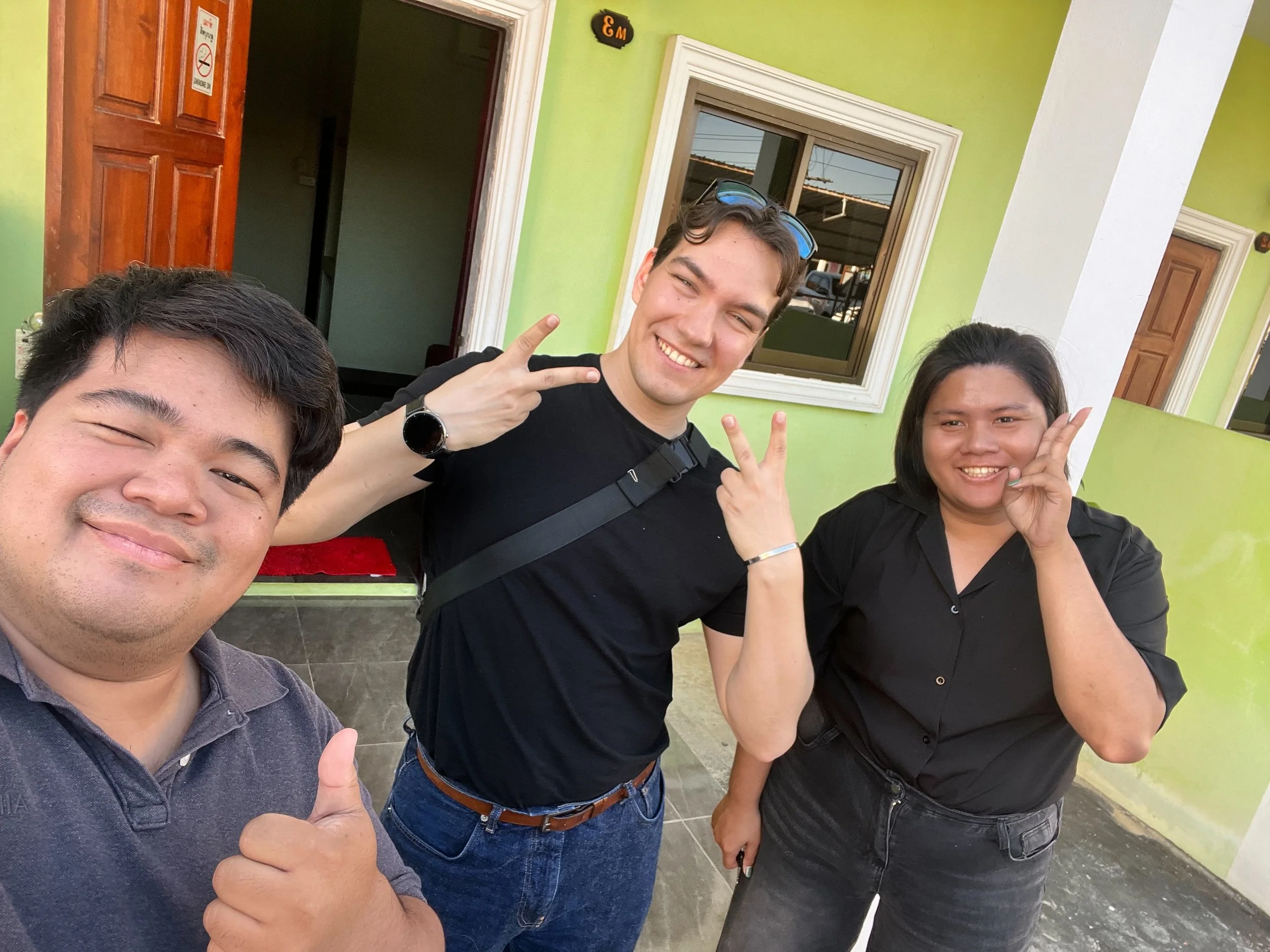 Three Kids English Thailand teachers smiling and posing for a selfie outside a building painted in green, with two of them making peace signs and one giving a thumbs up.