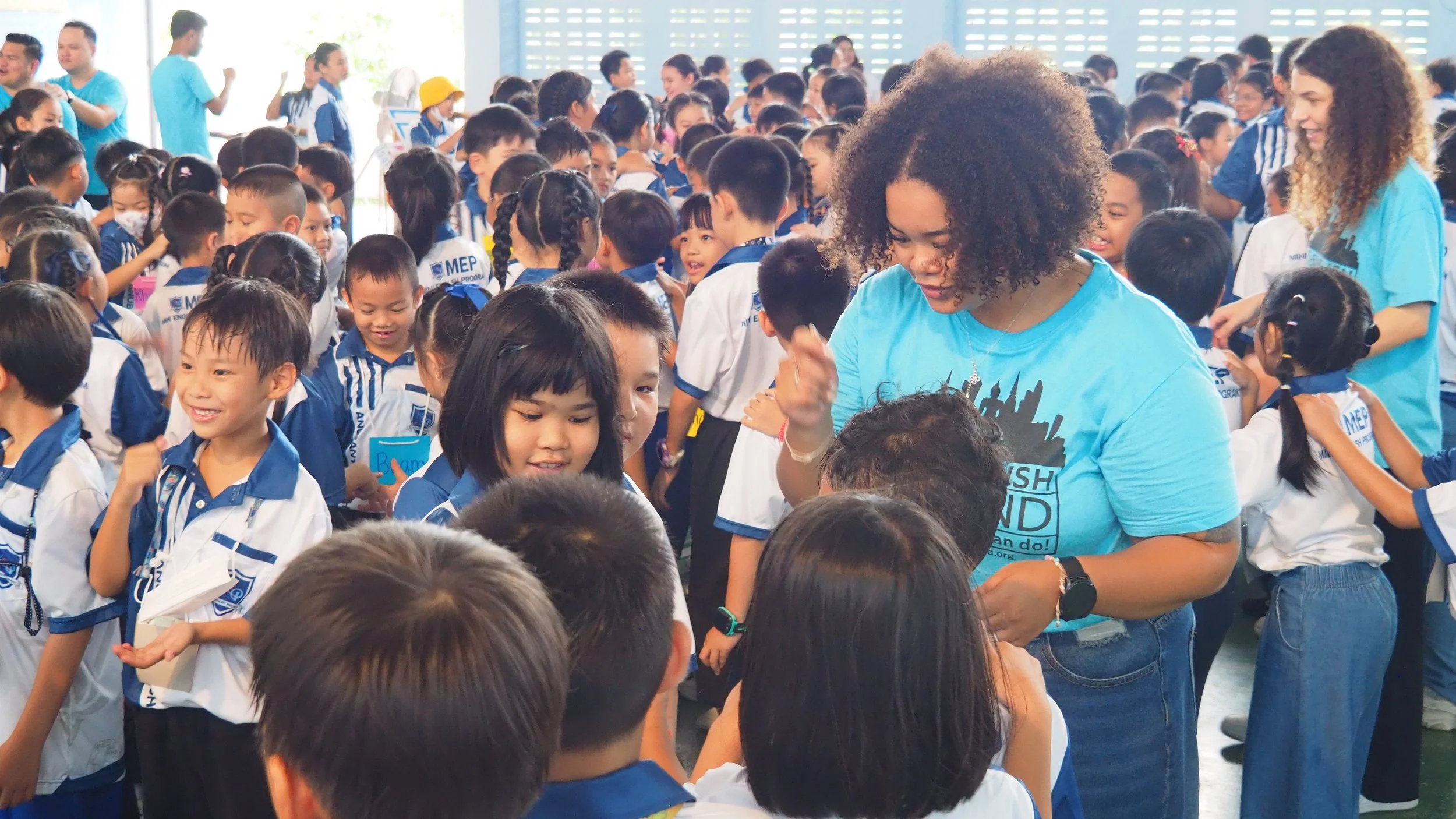 Group of children wearing school uniforms in a crowded indoor setting, with some Kids English Thailand teachers helping and engaging with them.