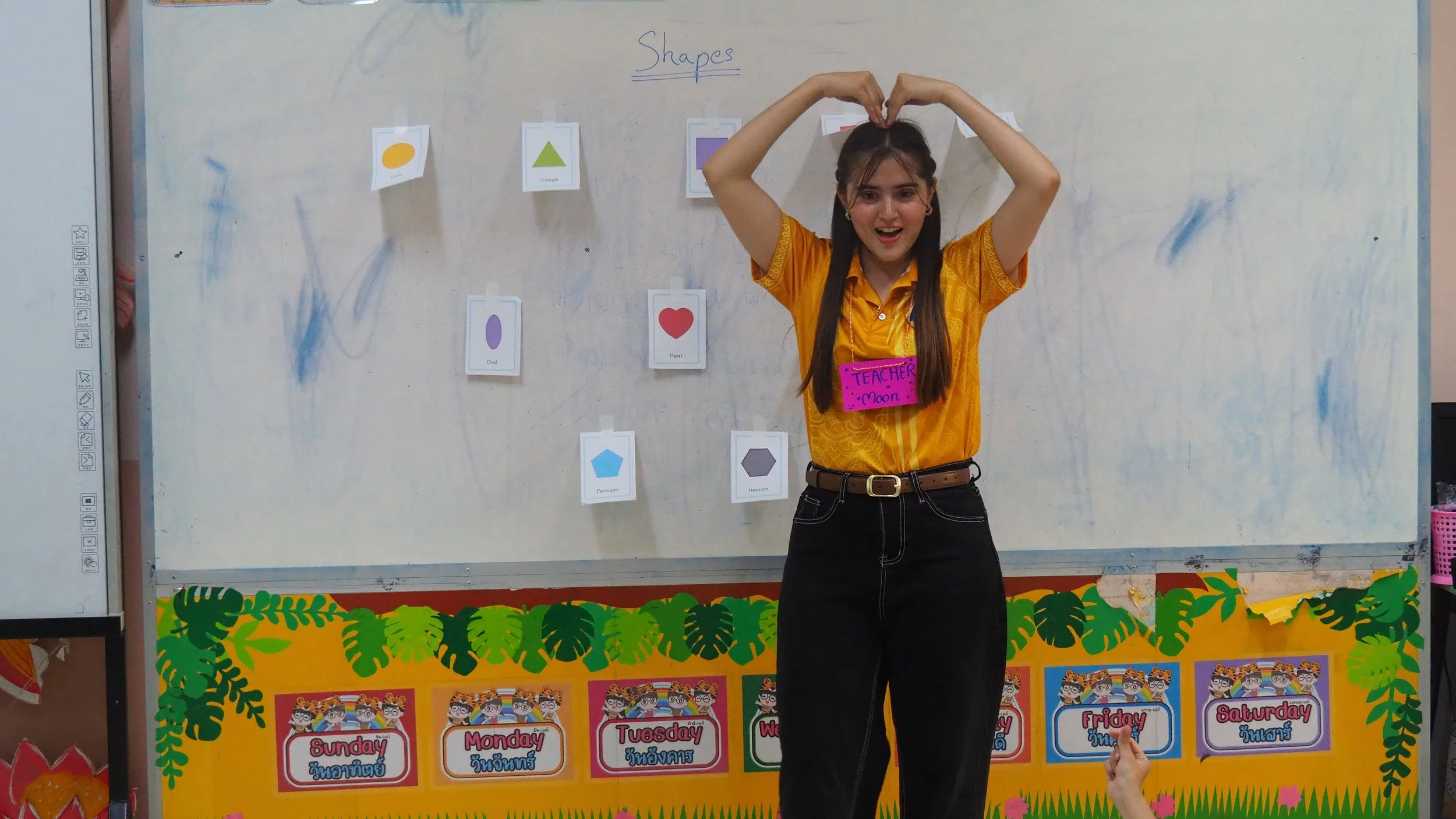 A young Kids English Thailand teacher with long brown hair, wearing a yellow shirt, stands in front of a classroom whiteboard. 