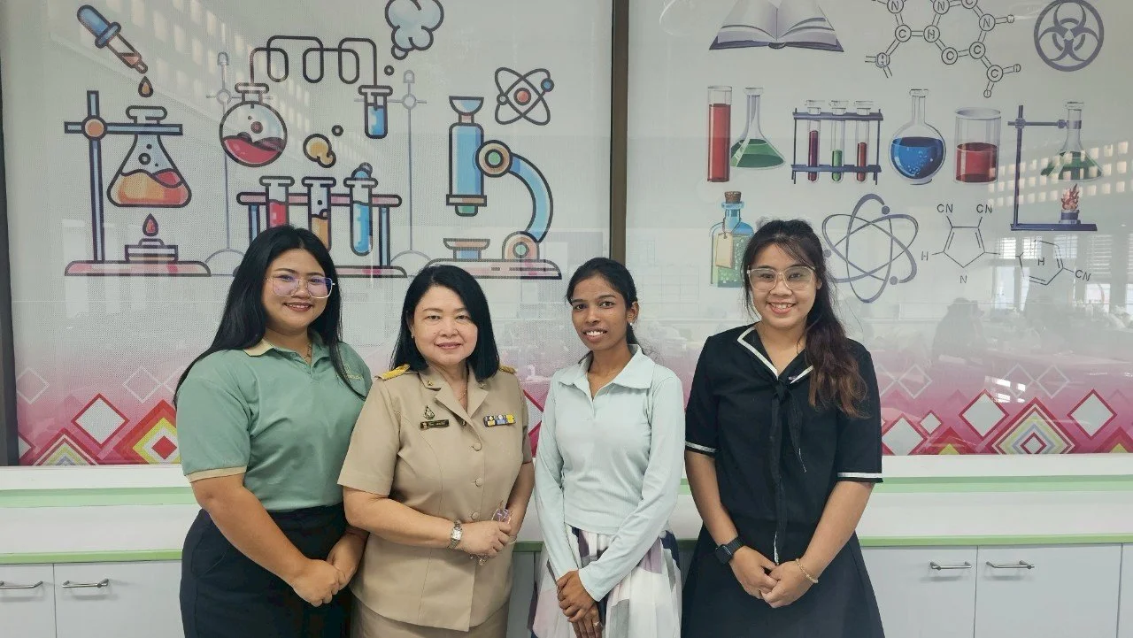 Four women from Kids English Thailand standing in front of a science-themed mural with laboratory glassware and chemical structures, smiling at the camera.