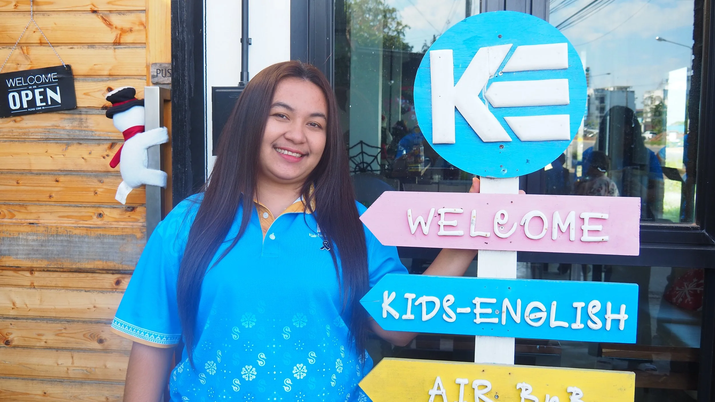 Smiling woman with long dark hair in a blue polo shirt standing outside a store with a wooden wall. She holds a sign with multiple arrows pointing in different directions, including a pink arrow labeled 'Welcome', a blue arrow labeled 'Kids-English', and a yellow arrow labeled 'Air Bob'. A black and white plush snowman hangs on the wall behind her, and an 'Open' sign is visible on the wall to her left.