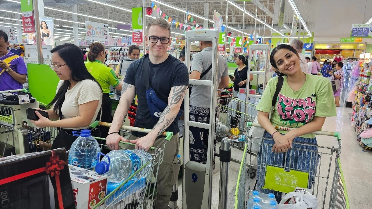 People from Kids English Thailand shopping in a brightly lit supermarket with checkout counters and shopping carts filled with water bottles and groceries.