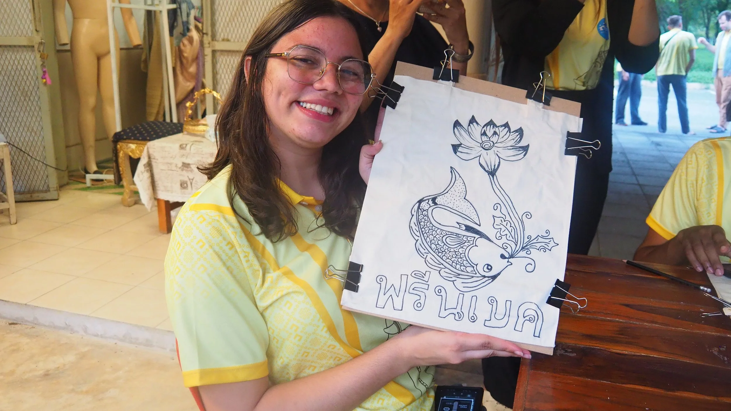 Smiling woman with glasses holding up a drawing of a fish and a lotus flower with Thai text, sitting at a table in a room with mannequins and other people.