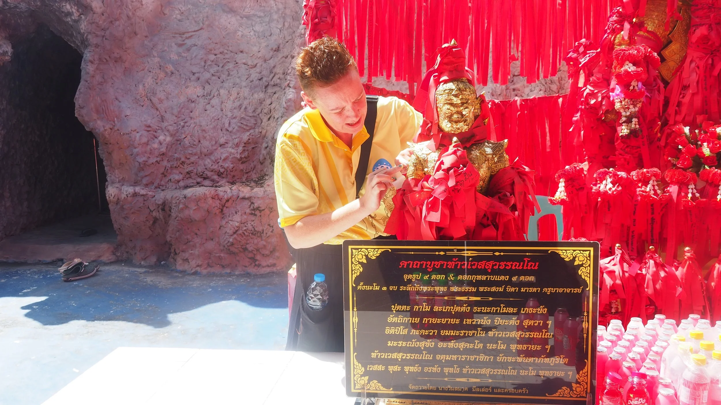 Kids English Thailand teacher in a yellow shirt placing a small object on a shrine with a golden Buddha statue, red ribbons, and offerings, with a black and gold sign and bottled water in front, against a rocky background.