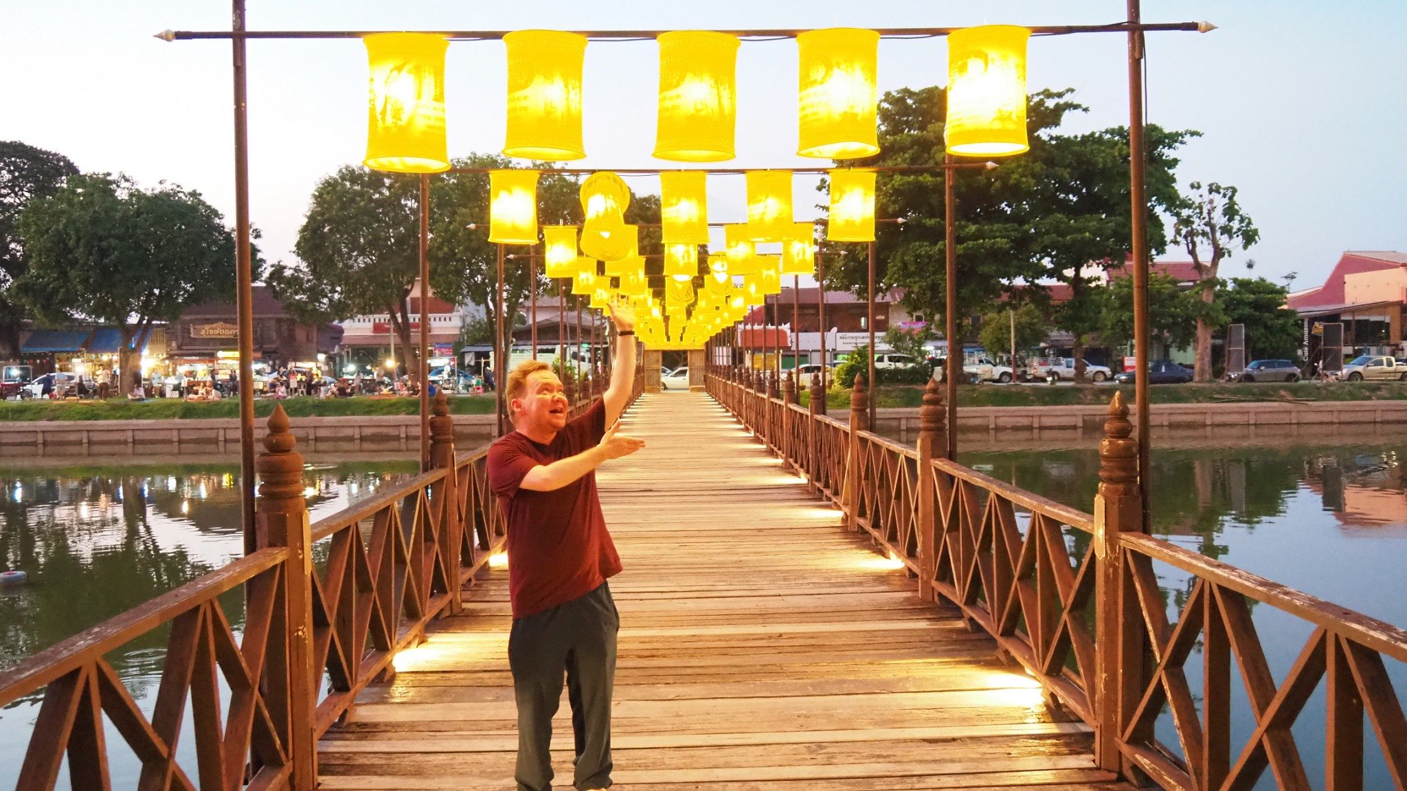 A Kids English Thailand teacher standing on a wooden bridge with yellow lanterns hanging above him at dusk, with a river and city buildings in the background.