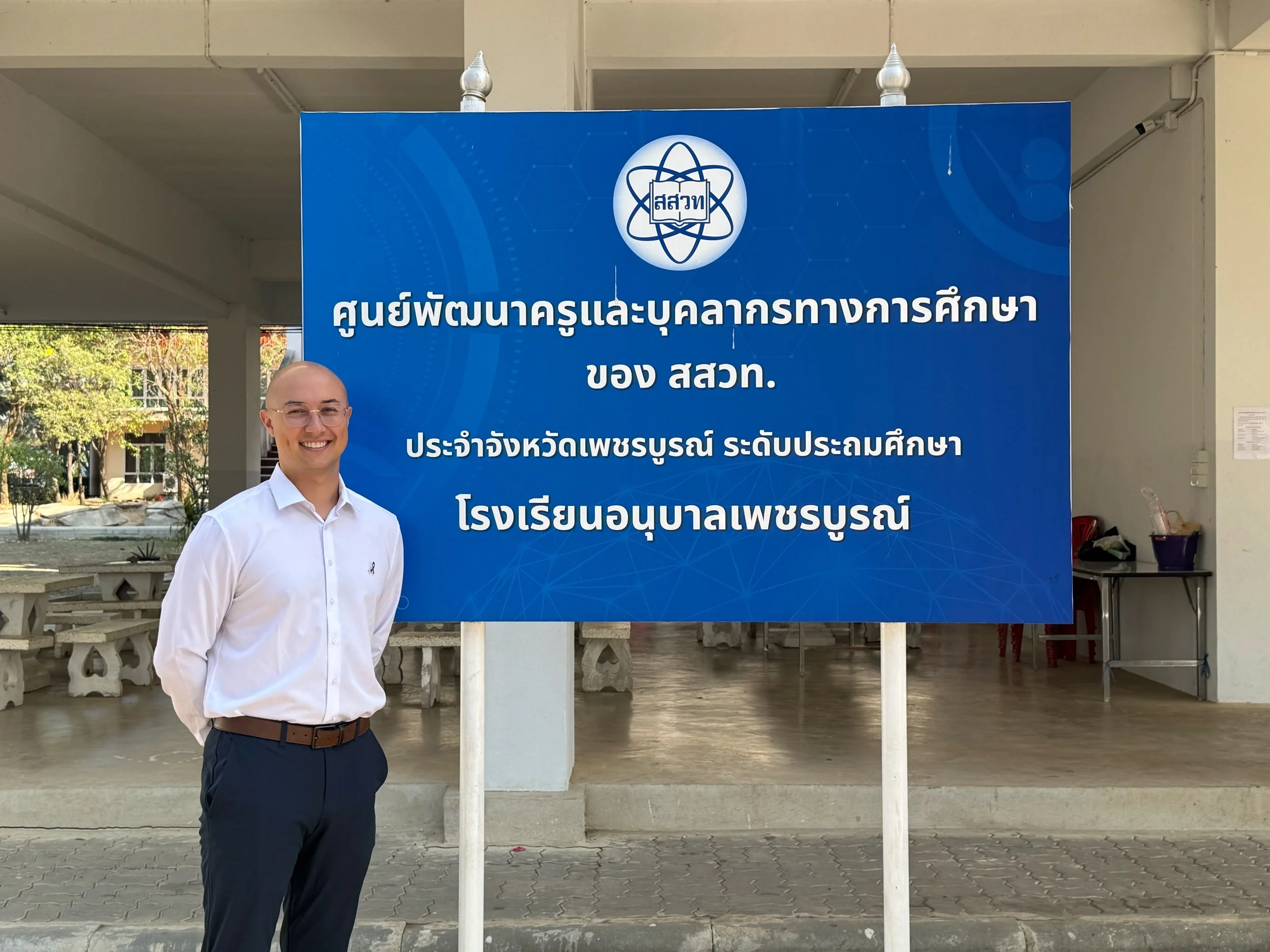 A smiling man from Kids English Thailand in a white shirt and dark pants standing next to a blue signboard with Thai text in front of a building.