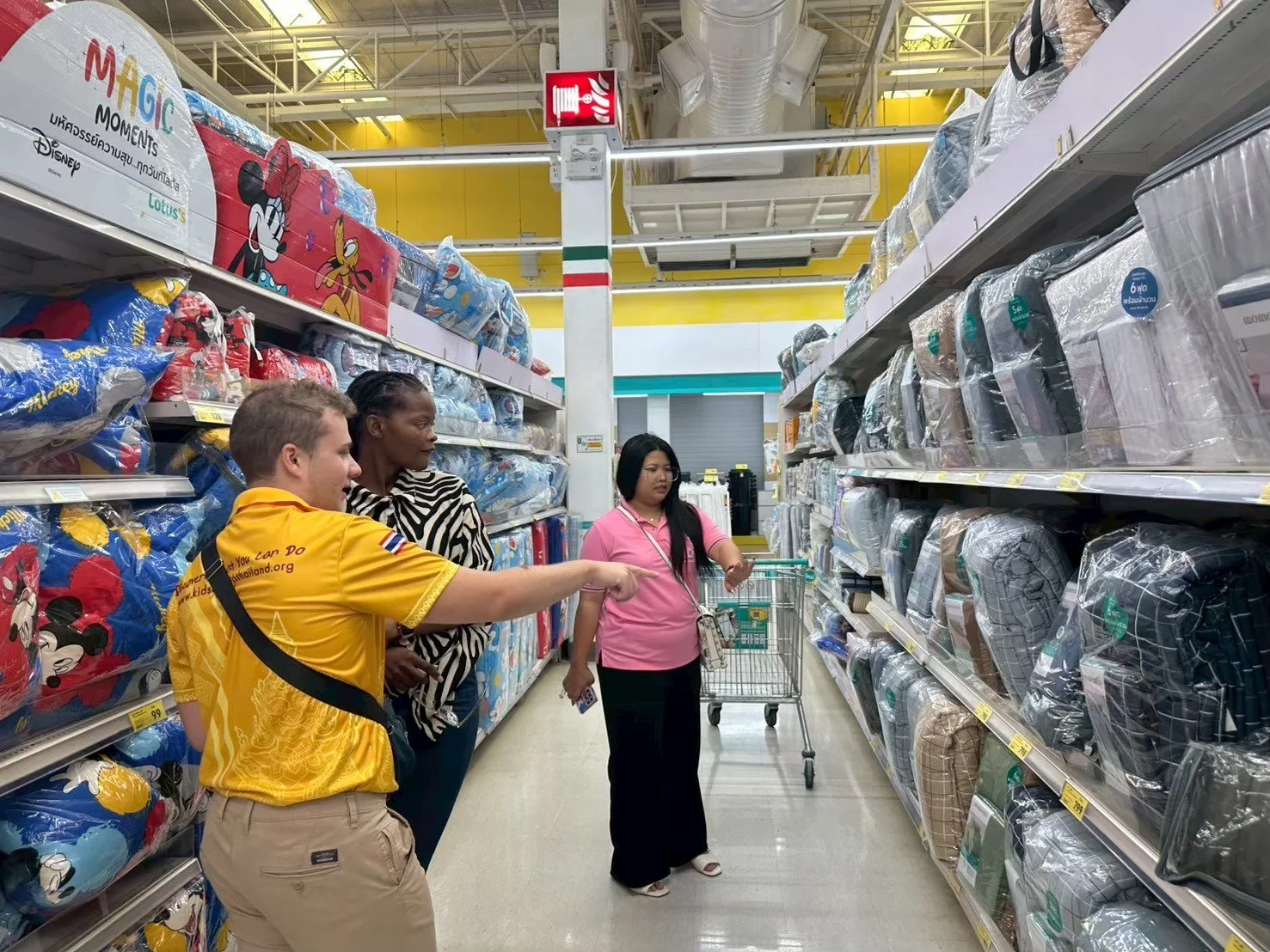 People from Kids English Thailand shopping for pillows in a store aisle with Disney-themed bedsheets on the left and cushions on the right.