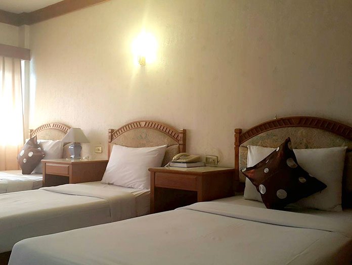 Three single beds with wooden headboards in a hotel room, each with pillows and nightstands, one with a lamp and a phone, a window with curtains on the left.