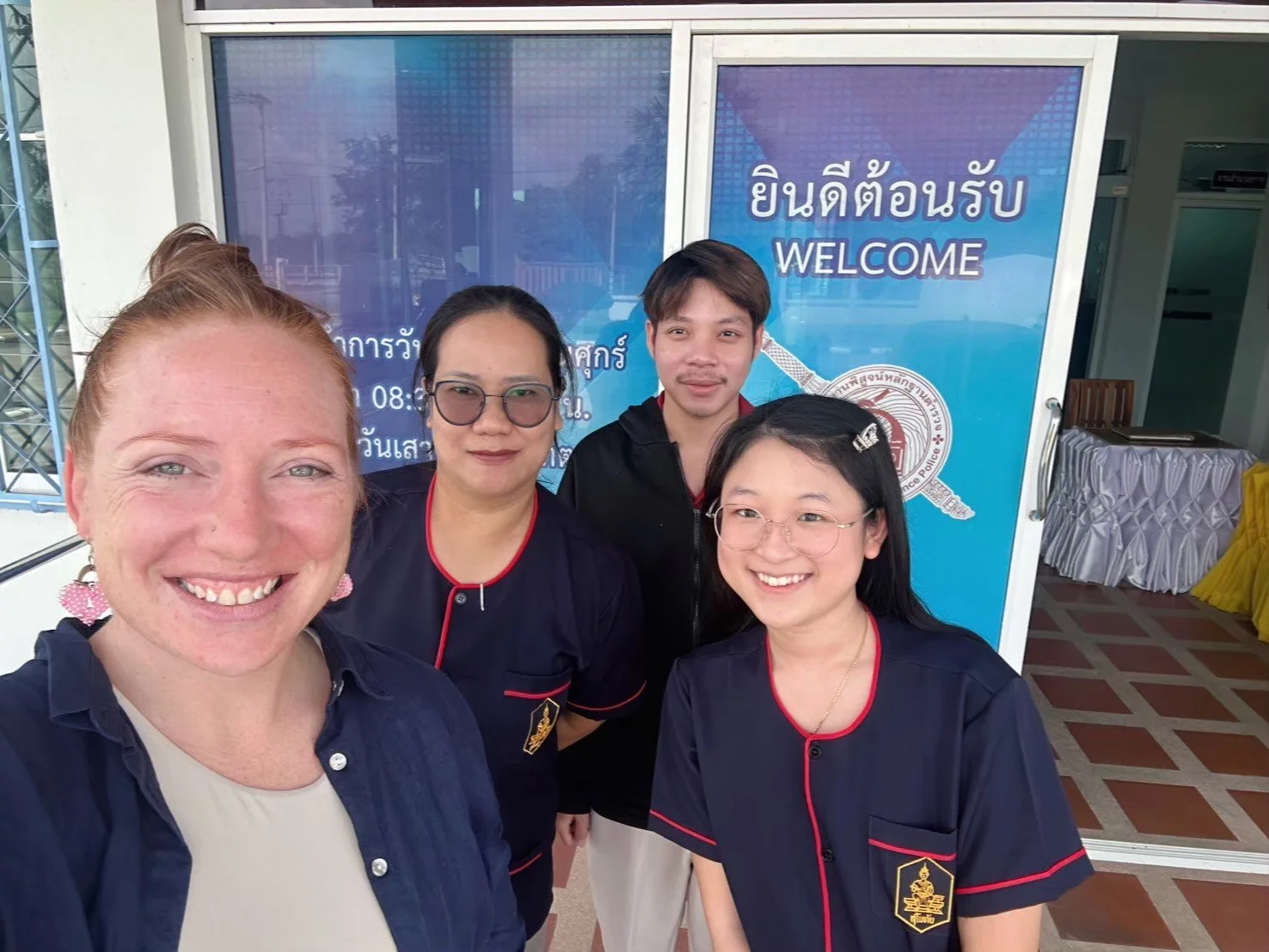 A group of five smiling diverse people from Kids English Thailand taking a selfie in front of a blue welcome sign with Thai and English text.