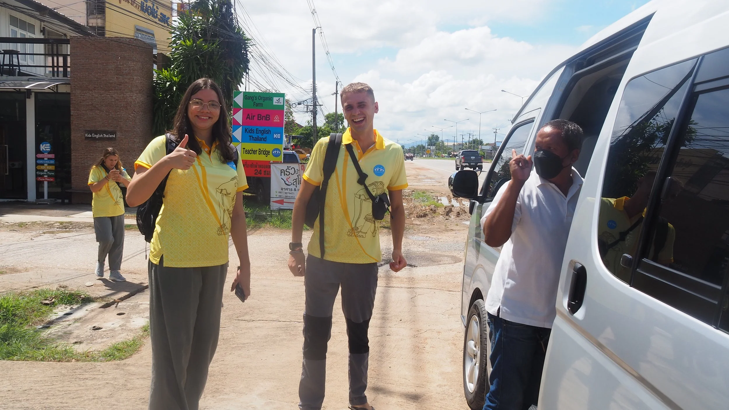 Three Kids English Thailand teachers standing outside on a sunny day. Two are wearing yellow shirts with logos, and one man is in a white shirt and black face mask next to a white vehicle.
