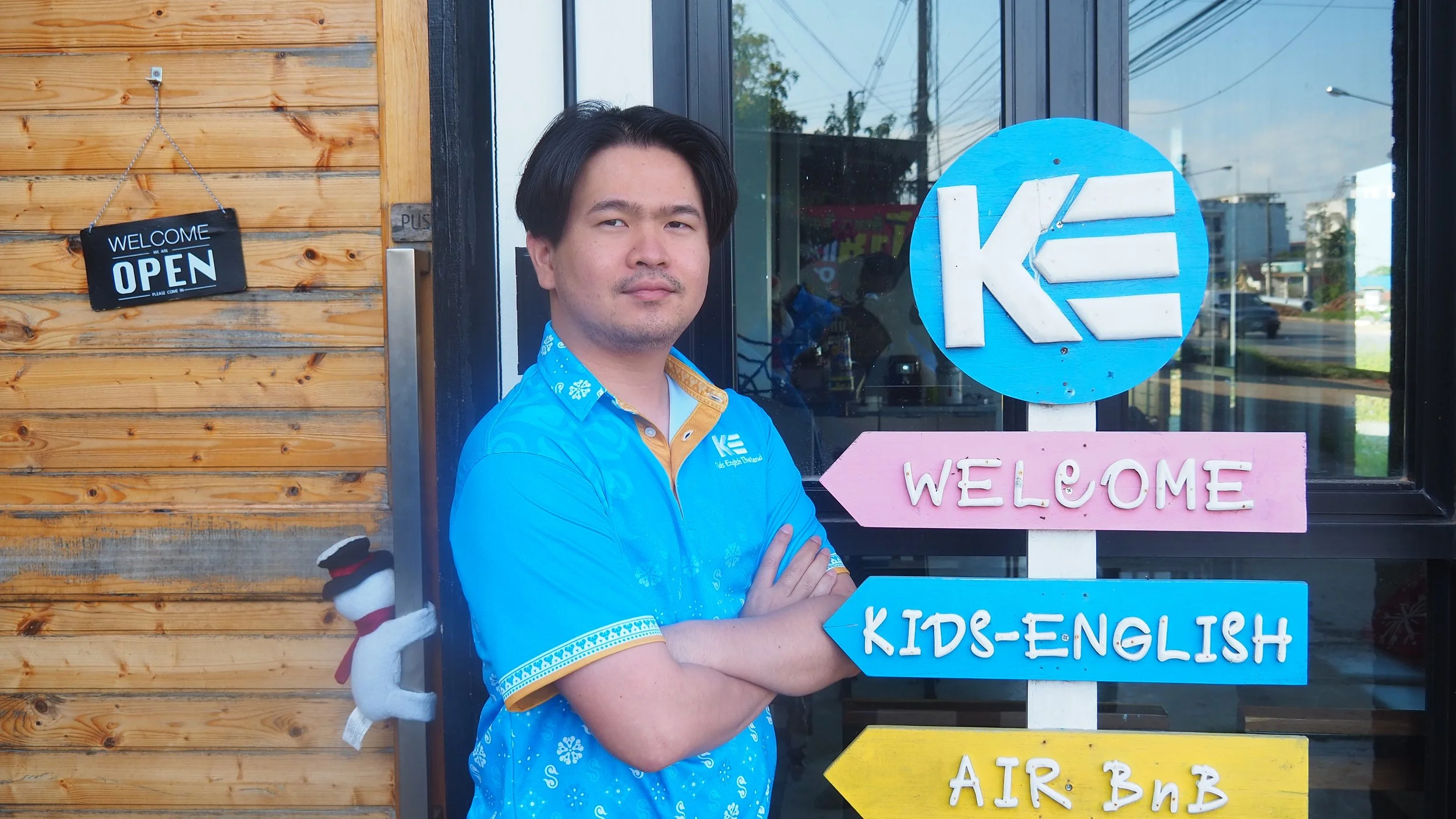 Man with crossed arms standing in front of a wooden building with signage that says 'Welcome,' 'Kids-English,' 'Air Bnb,' and 'K'