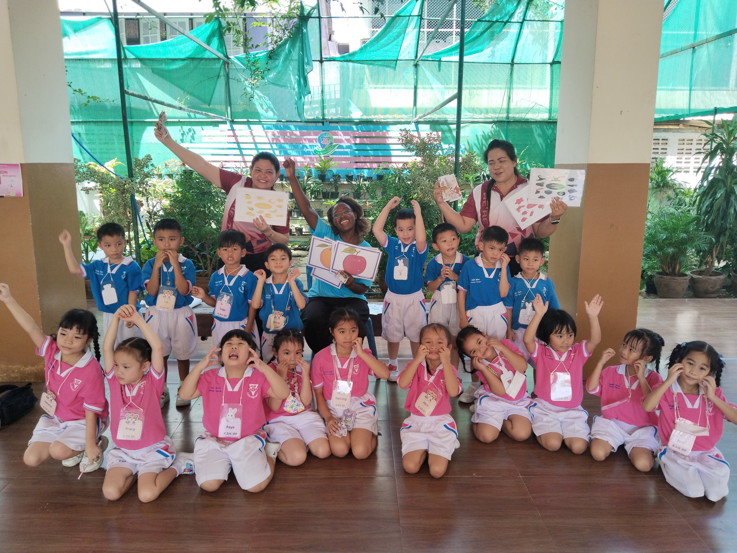 Group of young children and three Kids English Thailand teachers in a classroom with plants and green netting background, holding educational posters and posing with raised arms.