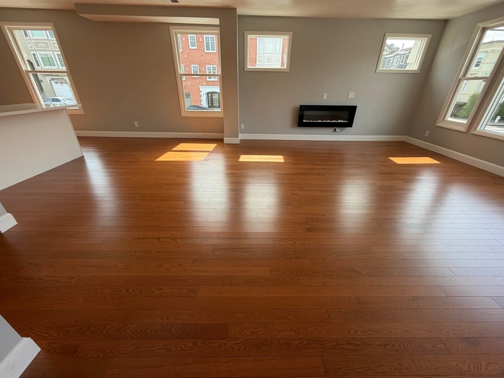 Empty living room with hardwood floors, multiple windows letting in sunlight, and a wall-mounted electric fireplace.