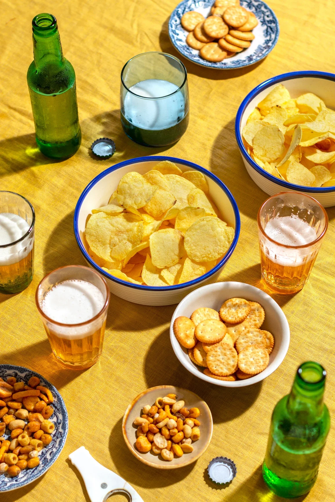 Varios platos y bebidas en una mesa con mantel amarillo. Incluye patatas fritas en tazones, galletas y galletas saladas, botellas de cerveza y vasos con distintas bebidas.